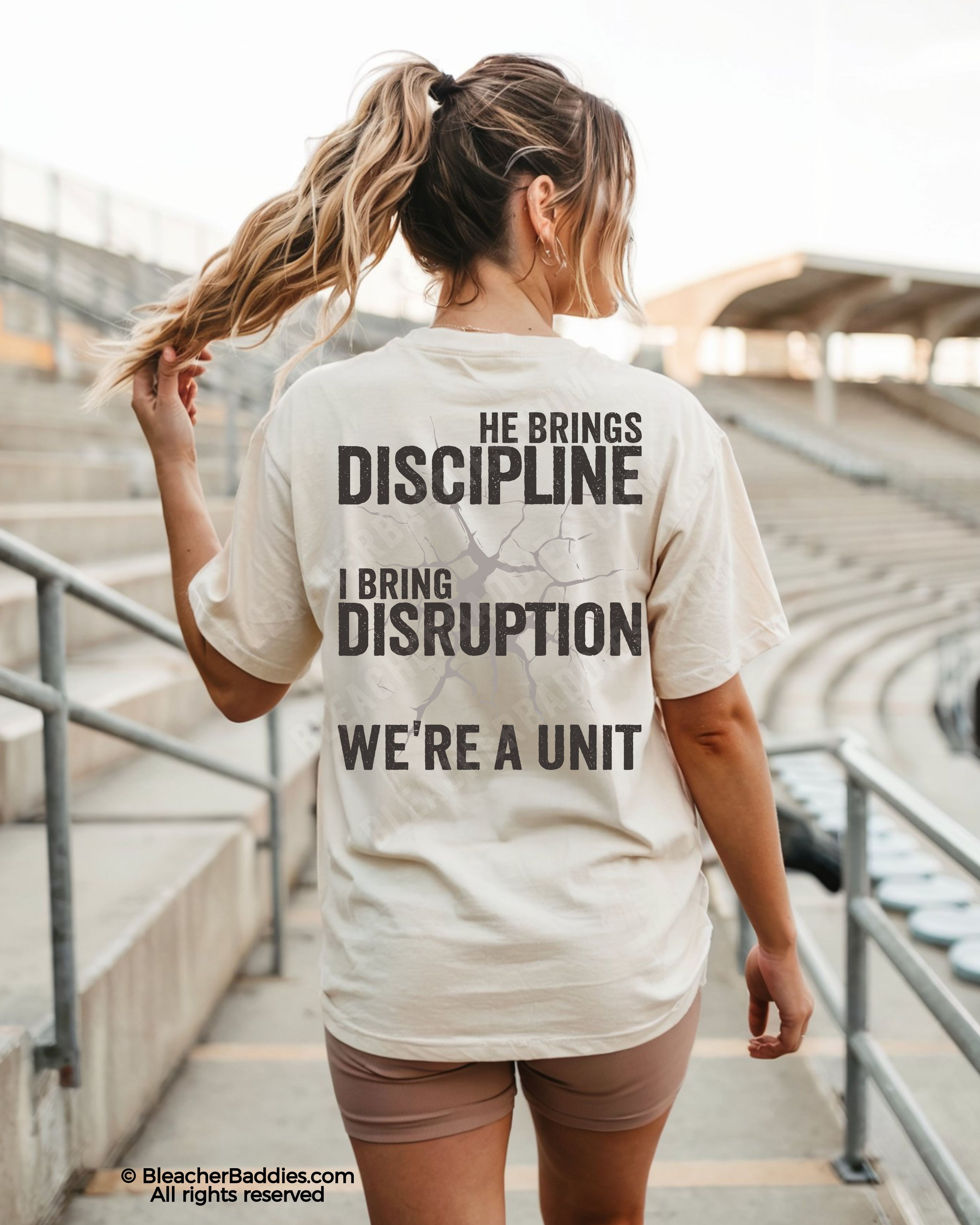 Back view of ivory Comfort Colors football mom game day shirt reading “He Brings Discipline I Bring Disruption We’re a Unit,” worn by woman walking through stadium seating