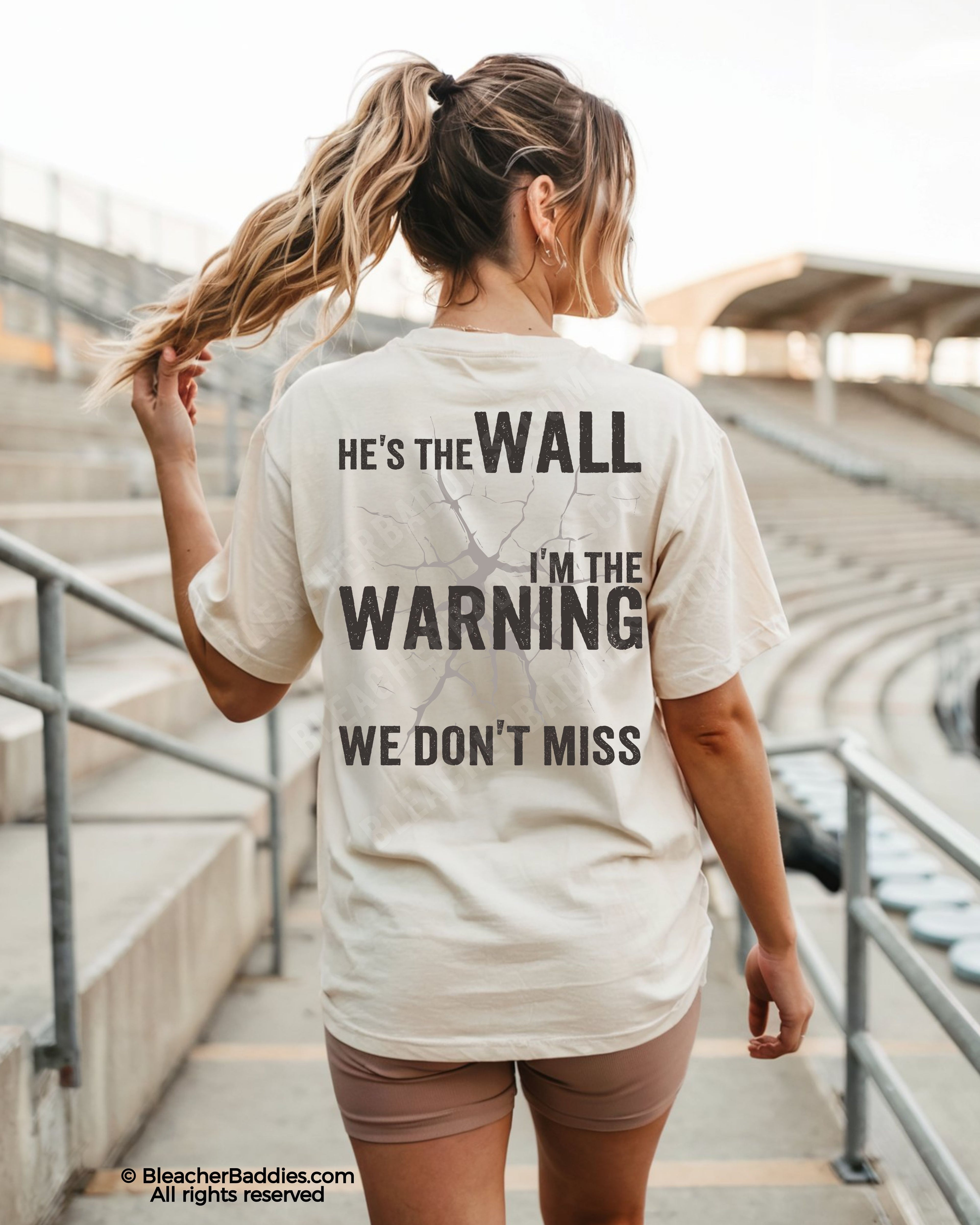 Woman wearing an ivory football mom game day shirt walking through stadium stands, featuring a large distressed black back graphic that reads “He’s the Wall I’m the Warning We Don’t Miss.”