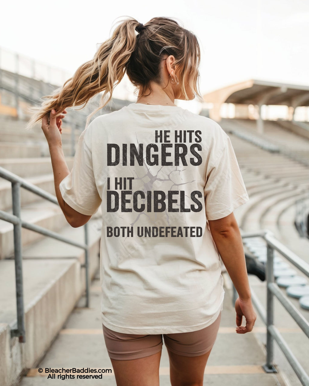 Woman wearing ivory Comfort Colors baseball mom shirt at stadium featuring bold distressed back graphic that says “He Hits Dingers I Hit Decibels Both Undefeated”