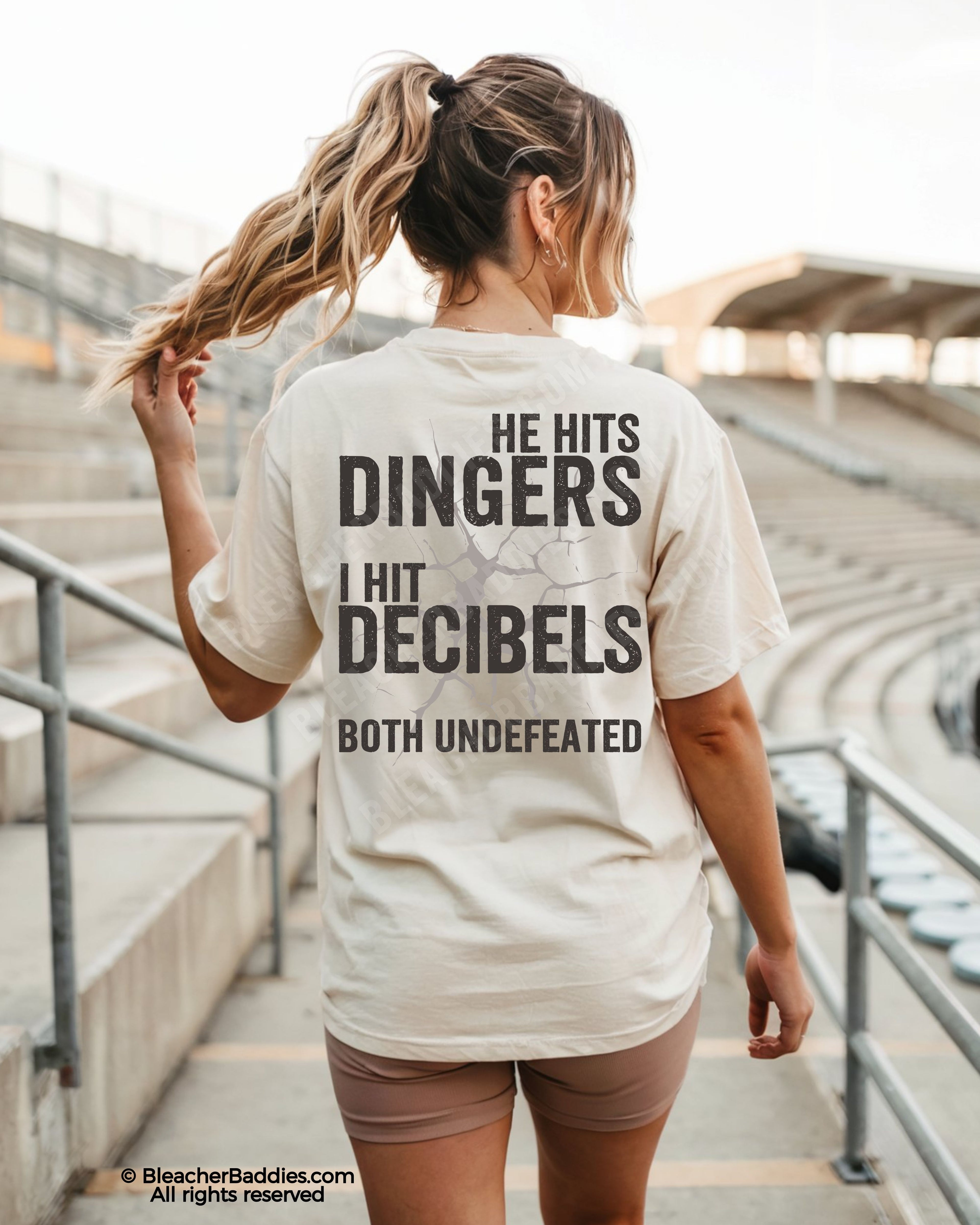Woman wearing ivory Comfort Colors baseball mom shirt at stadium featuring bold distressed back graphic that says “He Hits Dingers I Hit Decibels Both Undefeated”