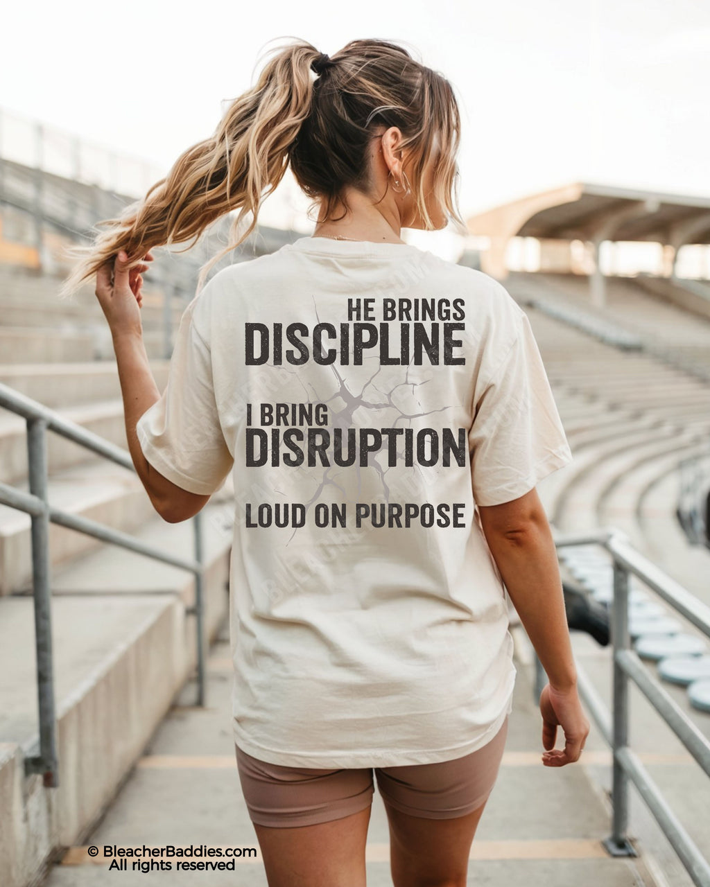 Woman wearing ivory Comfort Colors game day shirt at a baseball stadium featuring oversized distressed “He Brings Discipline I Bring Disruption” back print with “Loud On Purpose” text.