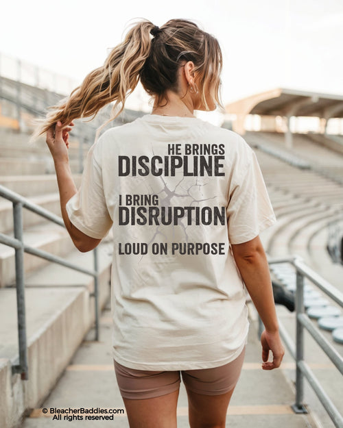 Woman wearing ivory Comfort Colors game day shirt at a baseball stadium featuring oversized distressed “He Brings Discipline I Bring Disruption” back print with “Loud On Purpose” text.