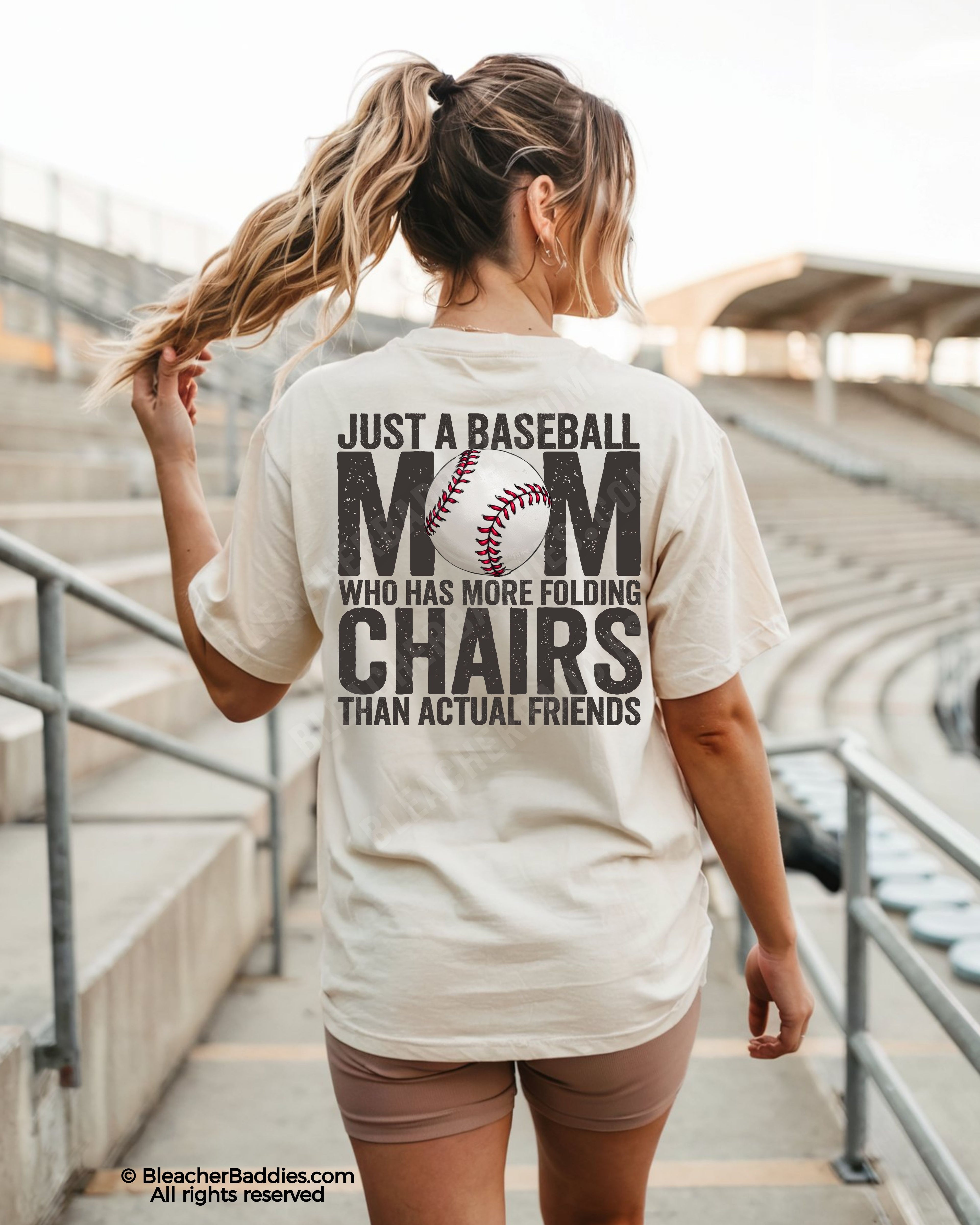Woman wearing ivory Comfort Colors baseball mom shirt in stadium with oversized distressed back graphic reading “Just a baseball mom who has more folding chairs than actual friends”