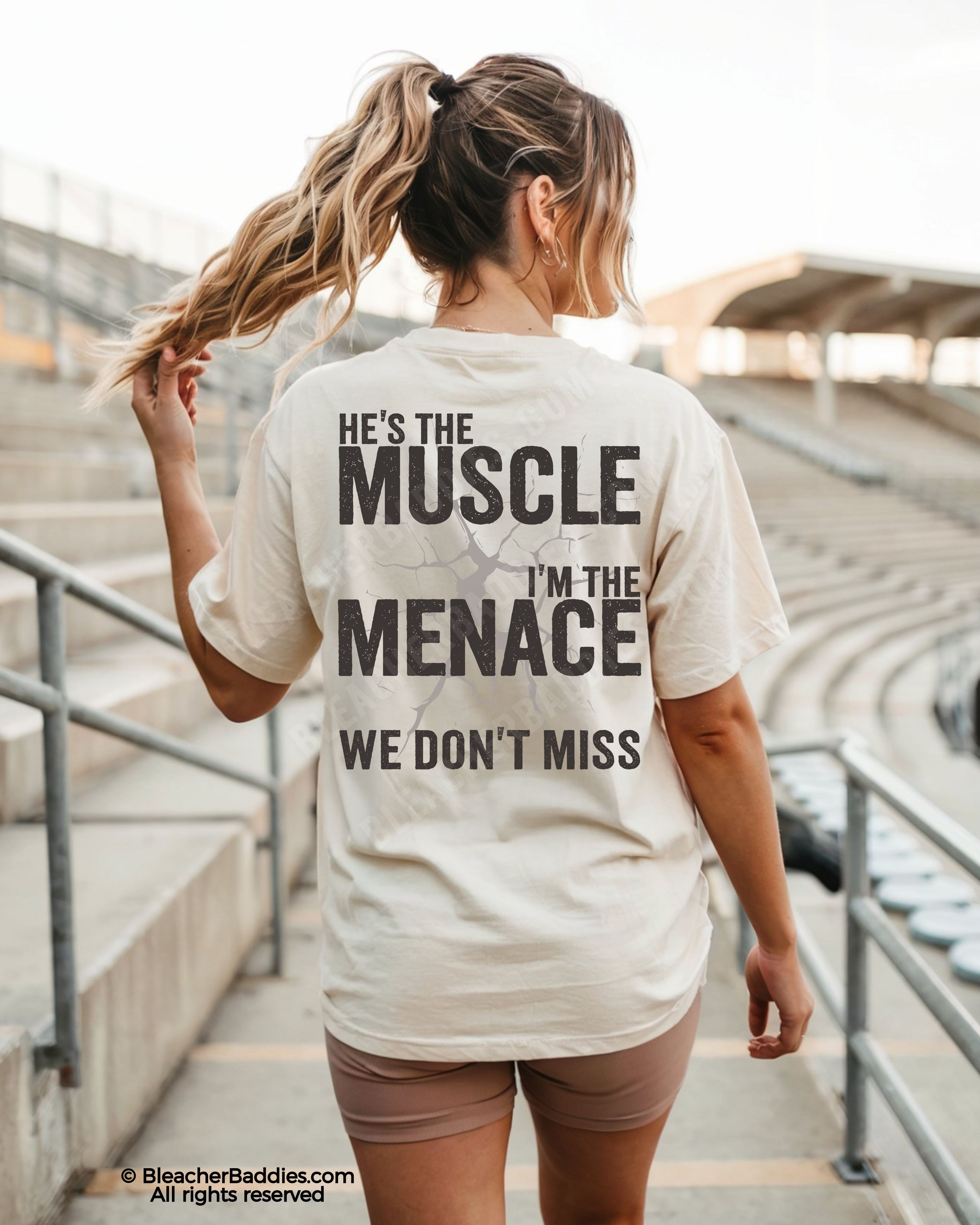 Woman wearing ivory Comfort Colors football mom game day shirt in stadium with back design that reads “He’s the muscle I’m the menace we don’t miss” in distressed black text with cracked graphic