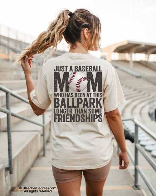 Woman walking through baseball stadium wearing ivory game day shirt with oversized back graphic reading “Just a Baseball Mom Who Has Been at This Ballpark Longer Than Some Friendships”