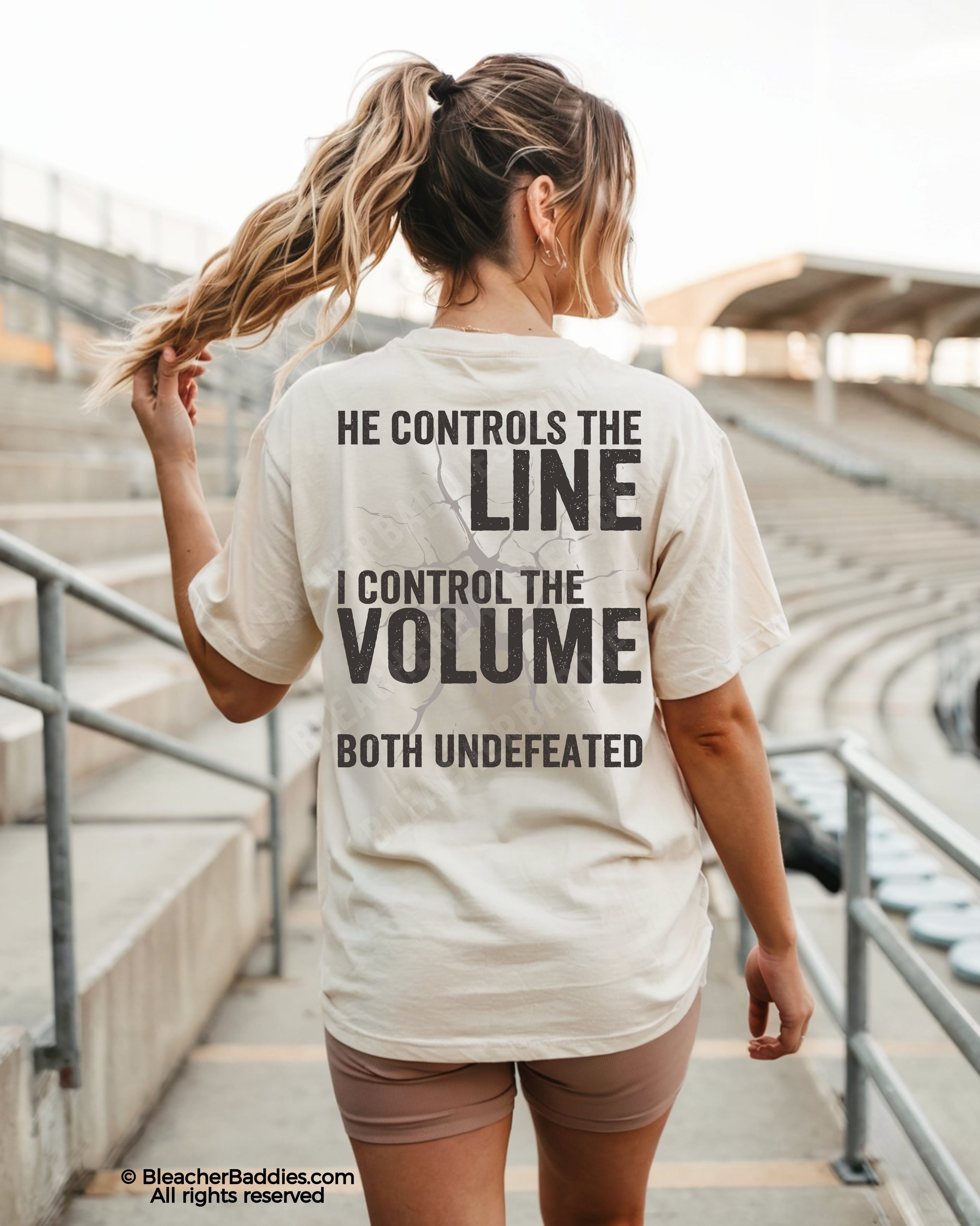 Back view of woman wearing ivory Comfort Colors football mom game day shirt in stadium featuring “He Controls the Line I Control the Volume Both Undefeated” bold graphic design