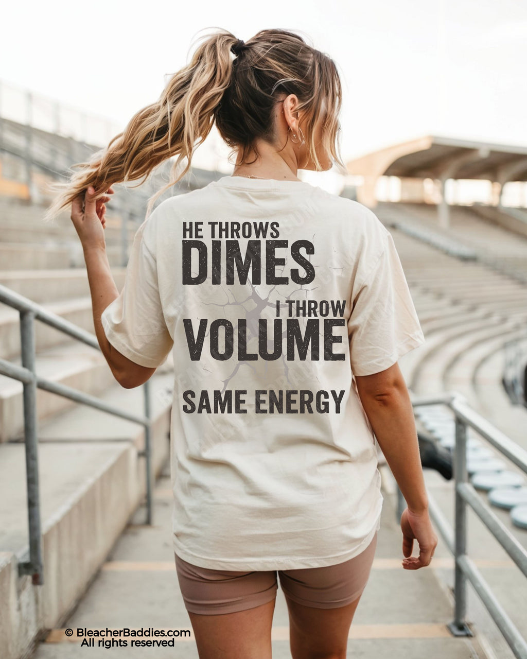Back view of ivory Comfort Colors game day shirt worn by woman in stadium, featuring bold text “He Throws Dimes I Throw Volume Same Energy” across the back.