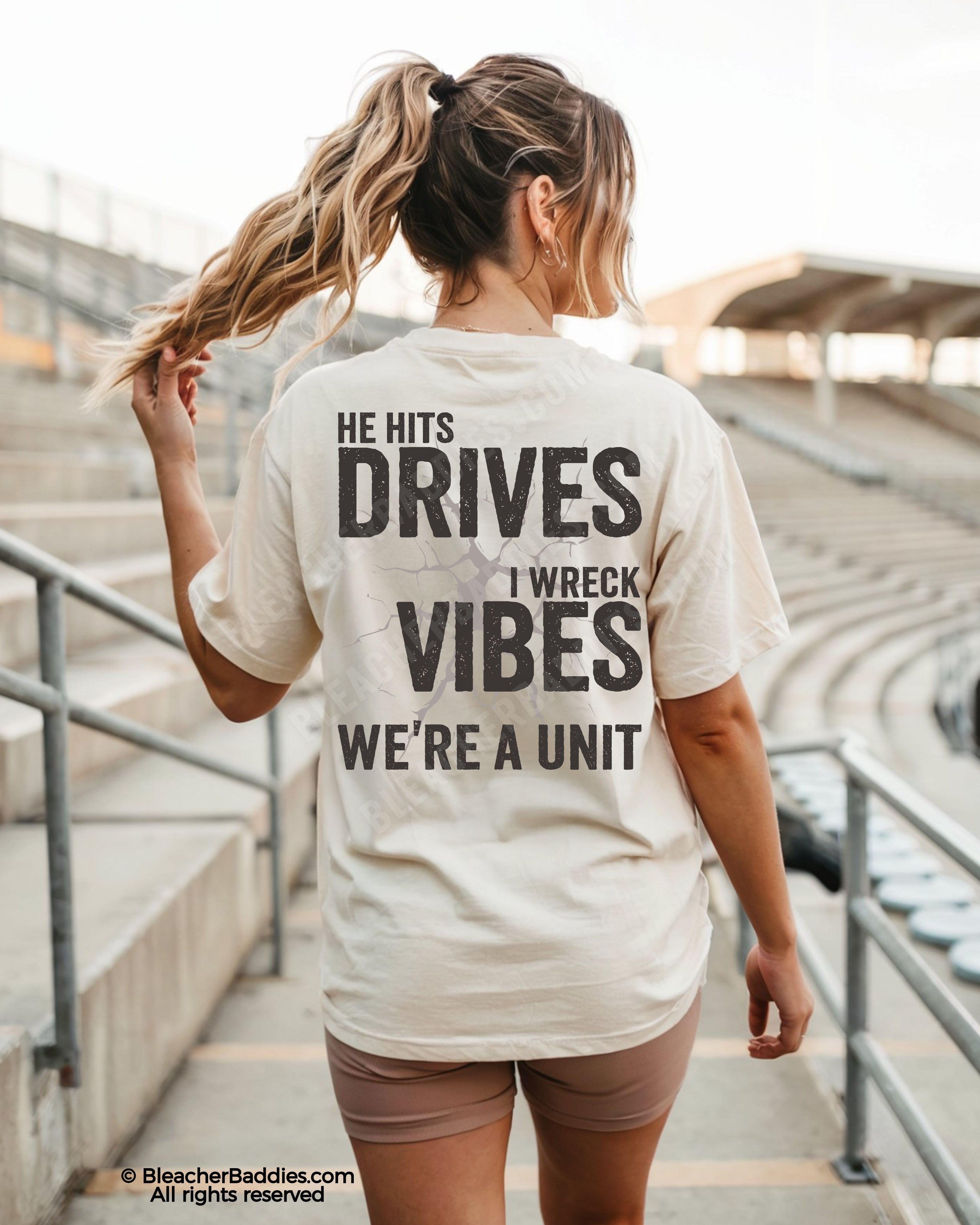 Woman wearing ivory Comfort Colors baseball mom game day shirt in stadium with oversized distressed back graphic reading “He Hits Drives I Wreck Vibes We’re A Unit” featuring cracked vintage effect typography.