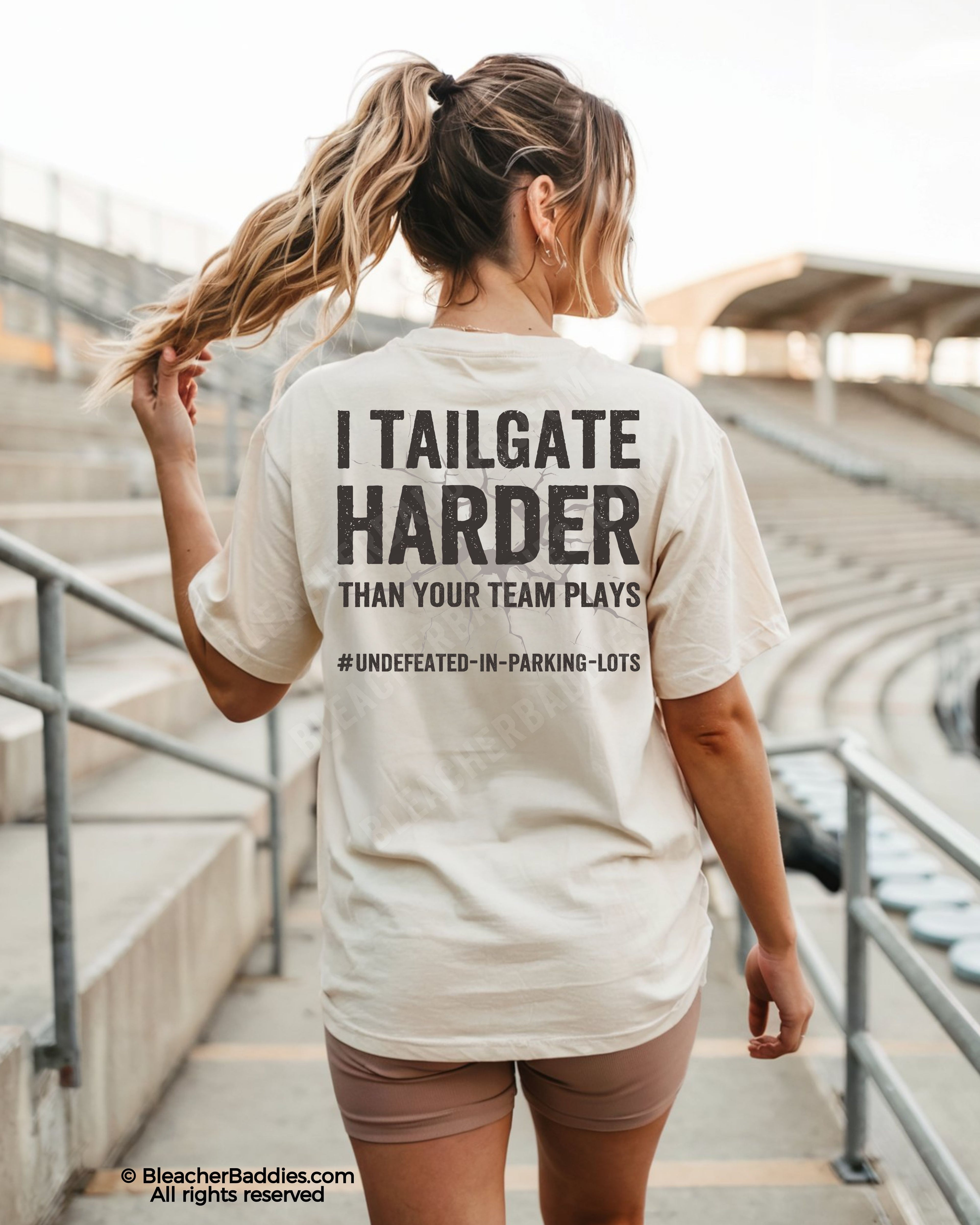 Woman wearing ivory Comfort Colors football mom game day shirt in stadium with back design that reads “I tailgate harder than your team plays #undefeated-in-parking-lots” in distressed black text with cracked graphic