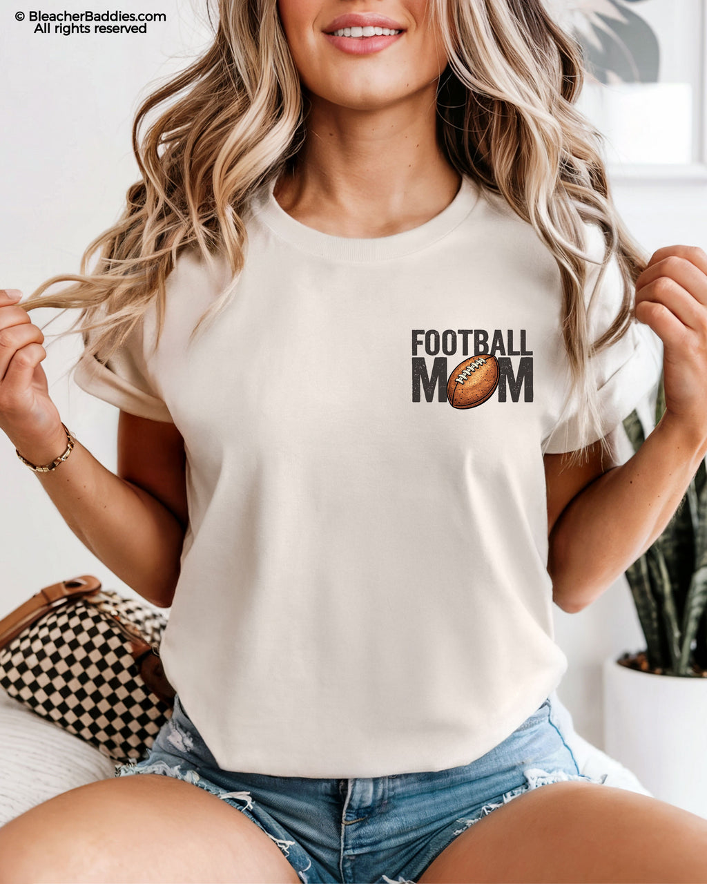 Woman wearing an ivory t-shirt with a small left chest graphic that reads “Football Mom” with a brown football replacing the “O” in mom, styled with a relaxed fit and paired with denim shorts.