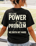 Back view of black Comfort Colors football mom game day shirt reading “He’s the Power I’m the Problem We Both Hit Hard,” worn by woman walking through stadium seating