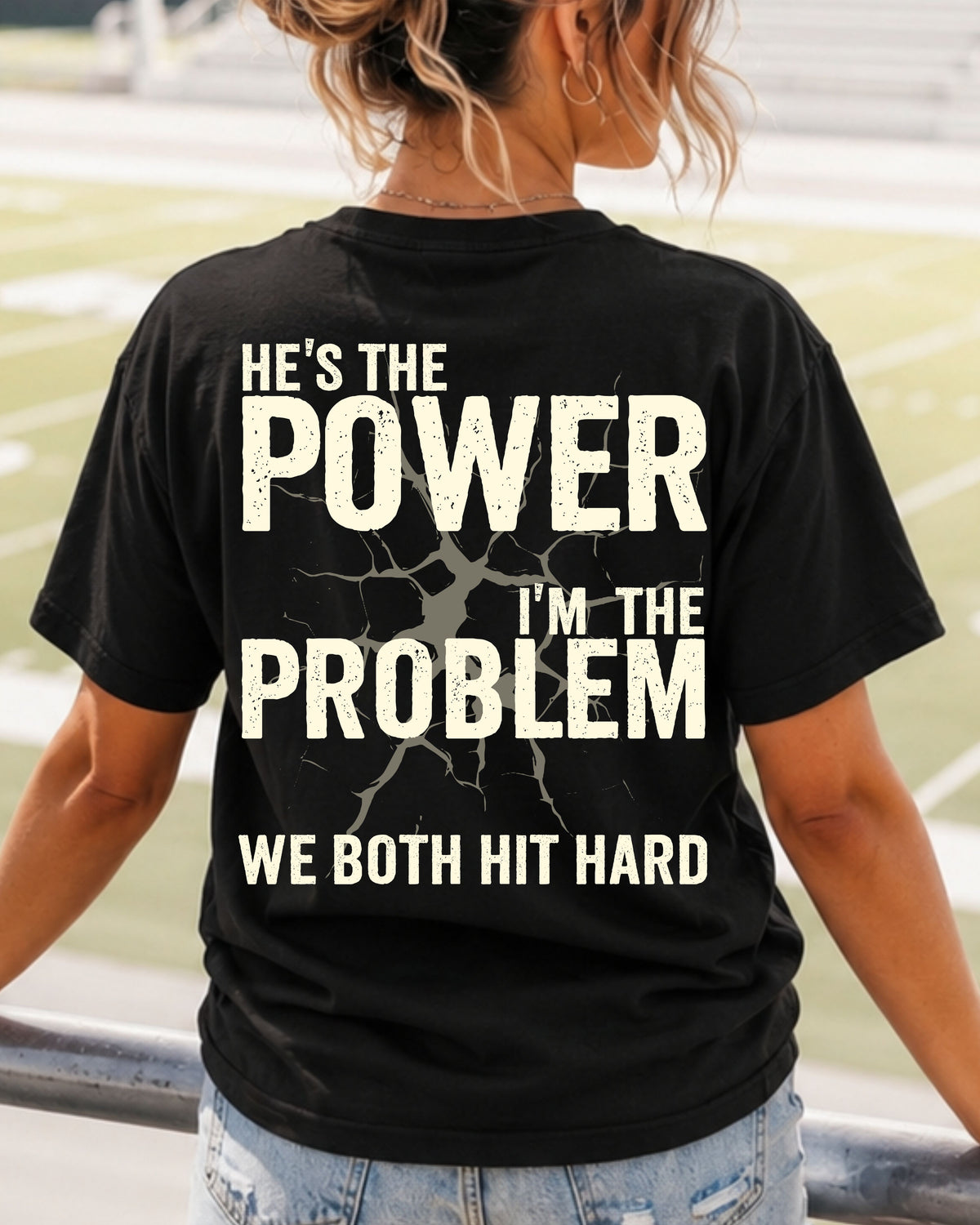Back view of black Comfort Colors football mom game day shirt reading “He’s the Power I’m the Problem We Both Hit Hard,” worn by woman walking through stadium seating