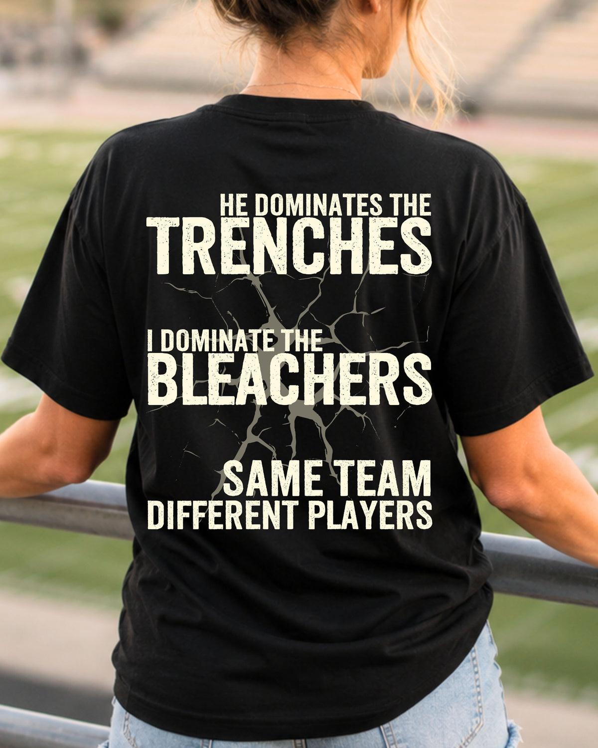 Woman wearing black comfort colors t-shirt in stadium showing back graphic that reads “He Dominates the Trenches I Dominate the Bleachers Same Team Different Players” Lineman mom shirt Offensive Lineman Shirt. Football Mom shirt.