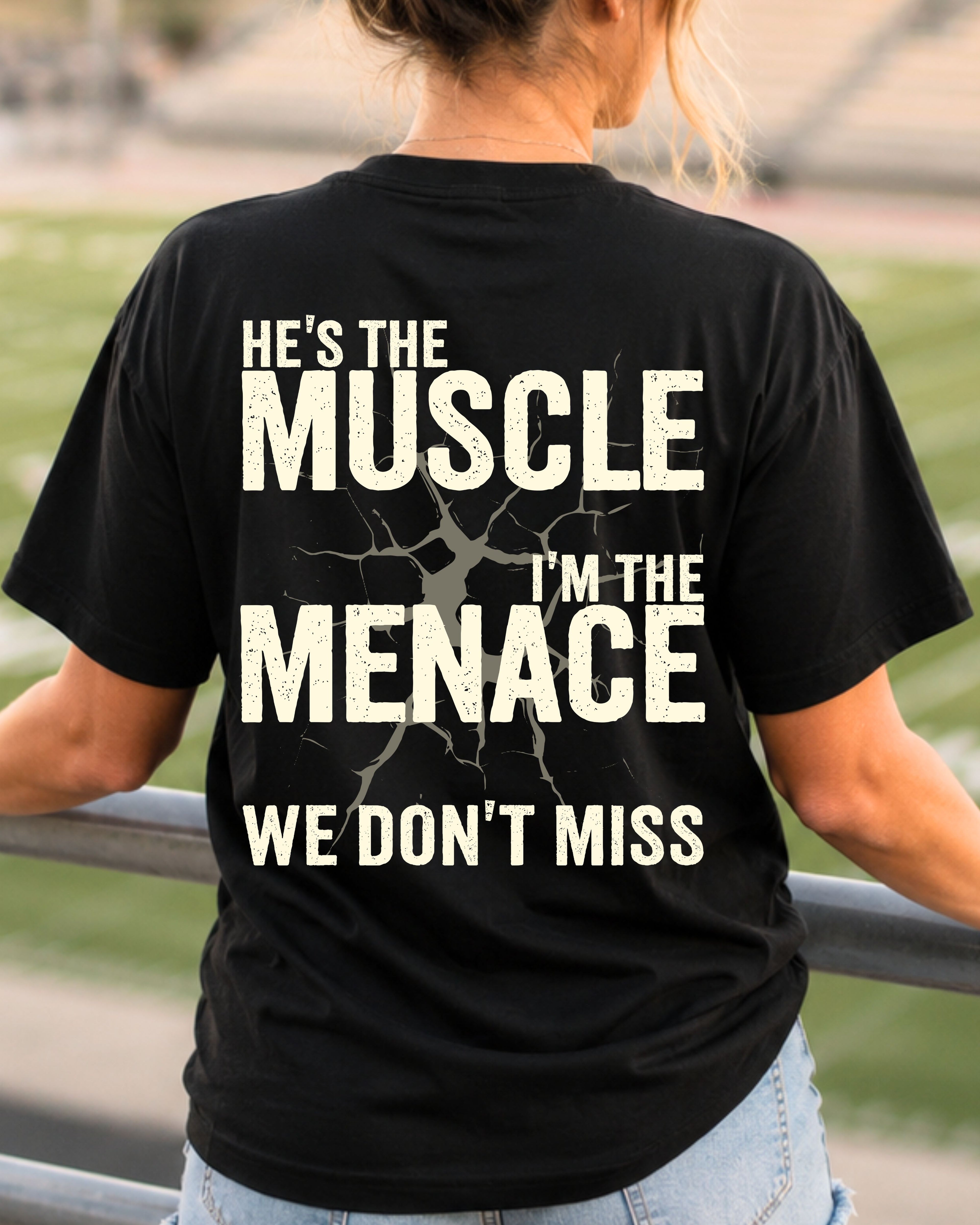 Woman wearing black Comfort Colors football mom game day shirt in stadium with back design that reads “He’s the muscle I’m the menace we don’t miss” in distressed cream text with cracked graphic.  Football mom shirt. Lineman mom shirt.