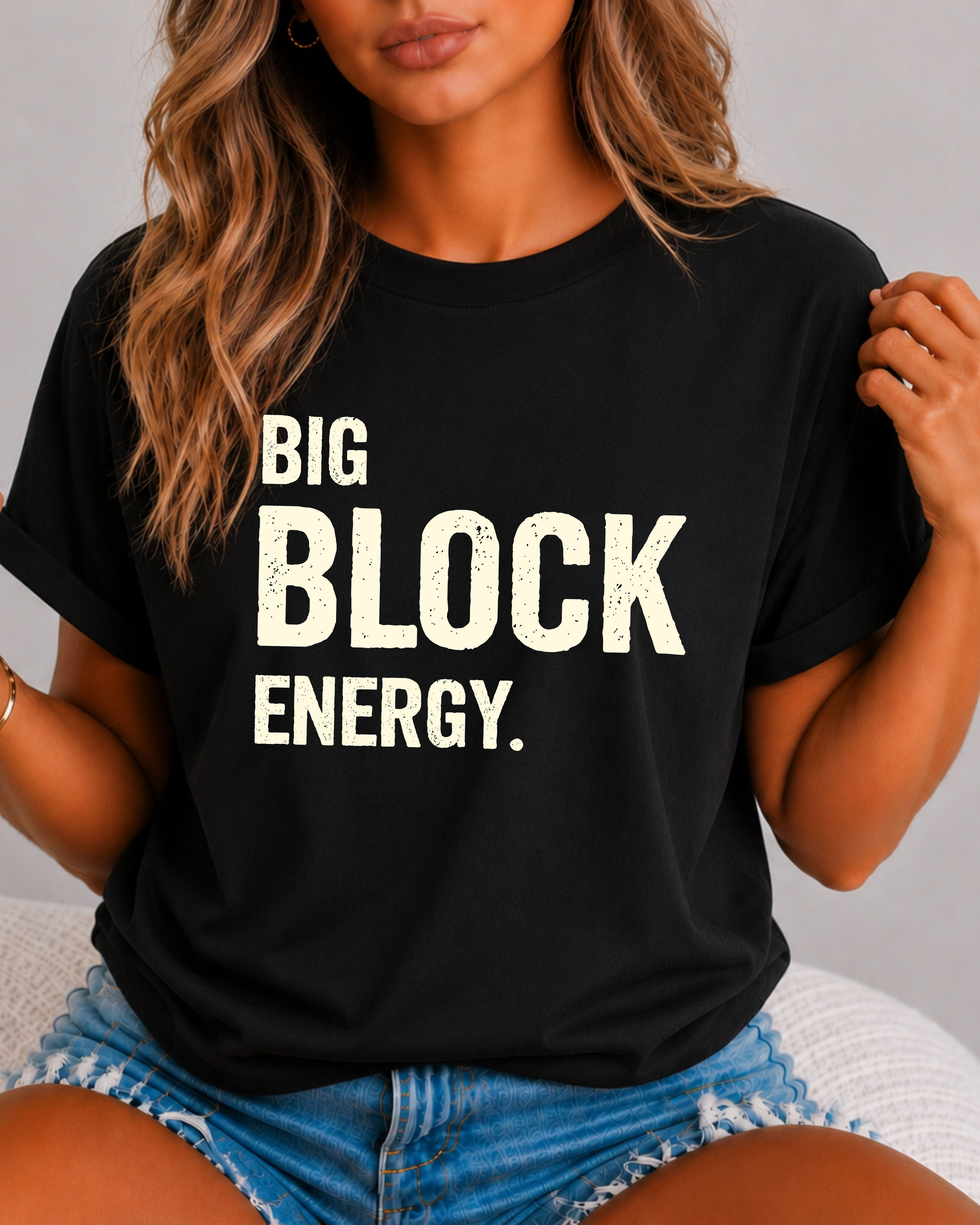woman wearing a black Comfort Colors t-shirt featuring the phrase "Big Block Energy" in distressed cream lettering. She is seated in light blue patterned shorts, making this the perfect football game day shirt for football moms, offensive line moms, lineman girlfriends, wives, and fans who love the big guys in the trenches.