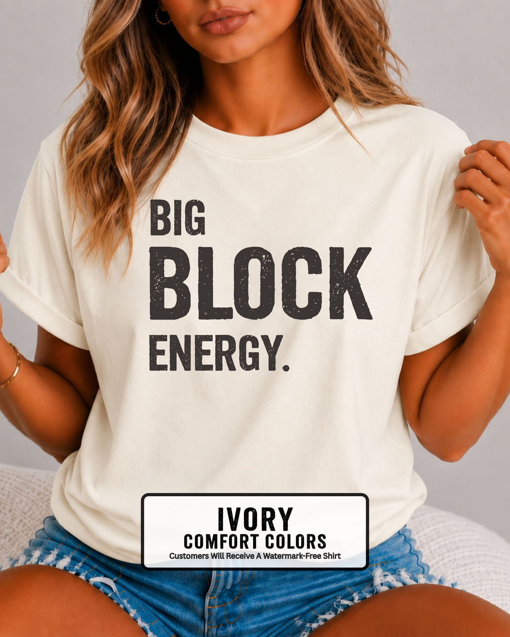 woman wearing an ivory Comfort Colors t-shirt featuring the phrase "Big Block Energy" in distressed dark lettering. She is seated in light blue patterned shorts, making this the perfect football game day shirt for football moms, offensive line moms, lineman girlfriends, wives, and fans who love the big guys in the trenches.