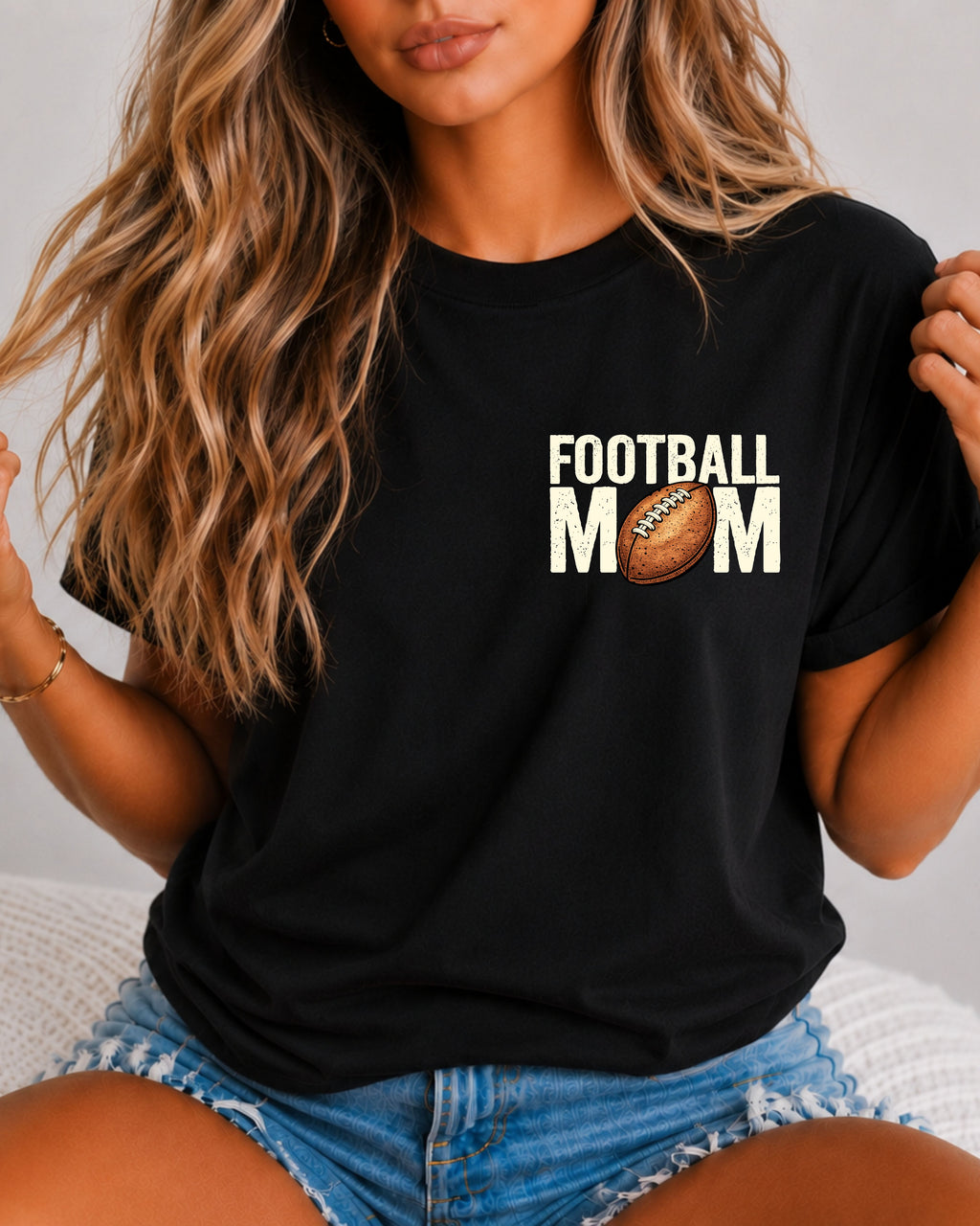 Woman wearing a black comfort colors t-shirt with a small left chest graphic that reads “Football Mom” with a brown football replacing the “O” in mom, styled with a relaxed fit and paired with denim shorts. Football Mom Shirt.