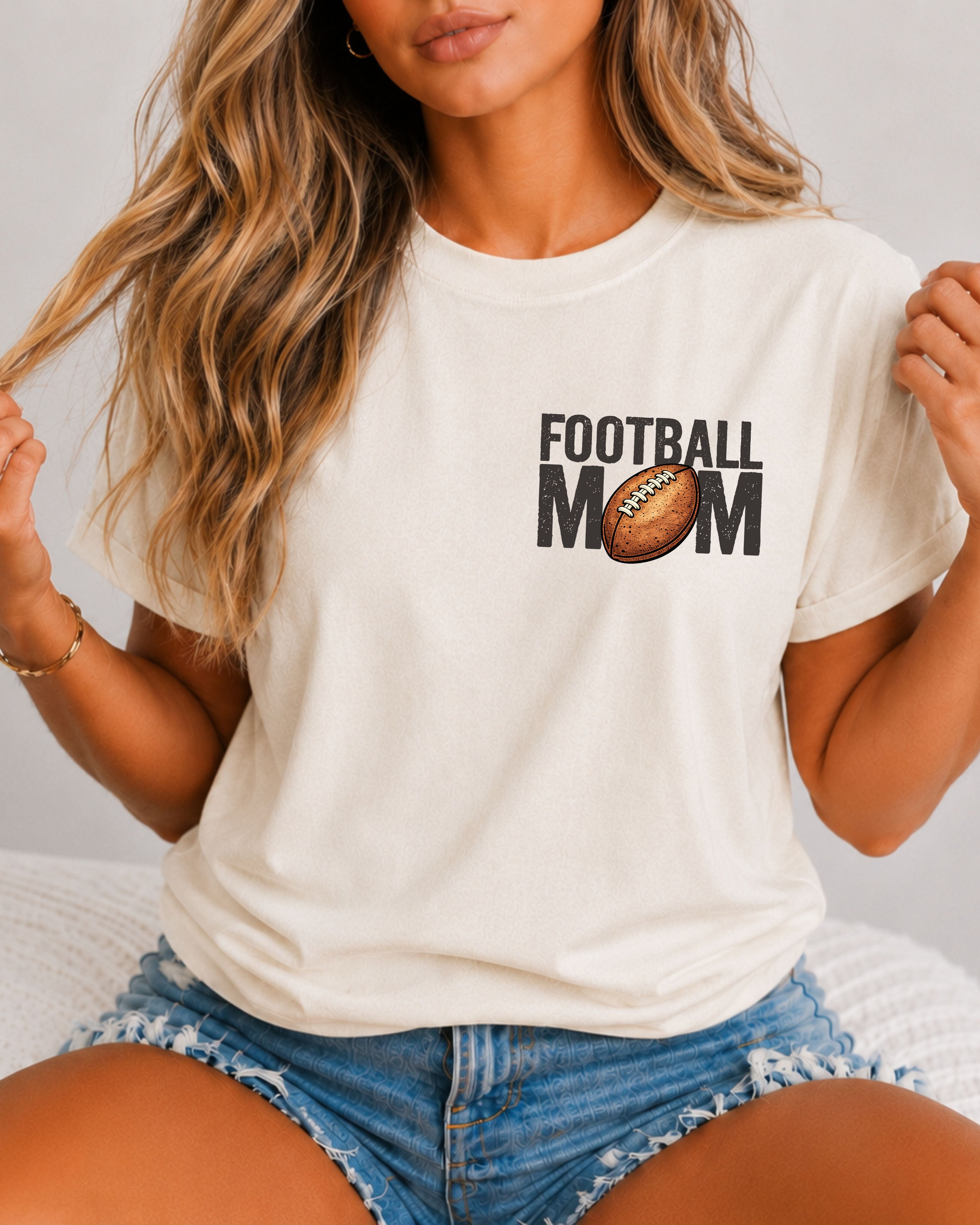 Woman wearing an ivory comfort colors t-shirt with a small left chest graphic that reads “Football Mom” with a brown football replacing the “O” in mom, styled with a relaxed fit and paired with denim shorts. Football Mom Shirt.