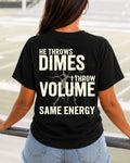 A woman seen from behind wearing an black comfort colors shirt with bold cream text that reads 'HE THROWS DIMES I THROW VOLUME SAME ENERGY' while standing in an outdoor stadium. Quarterback mom shirt. Football mom shirt.