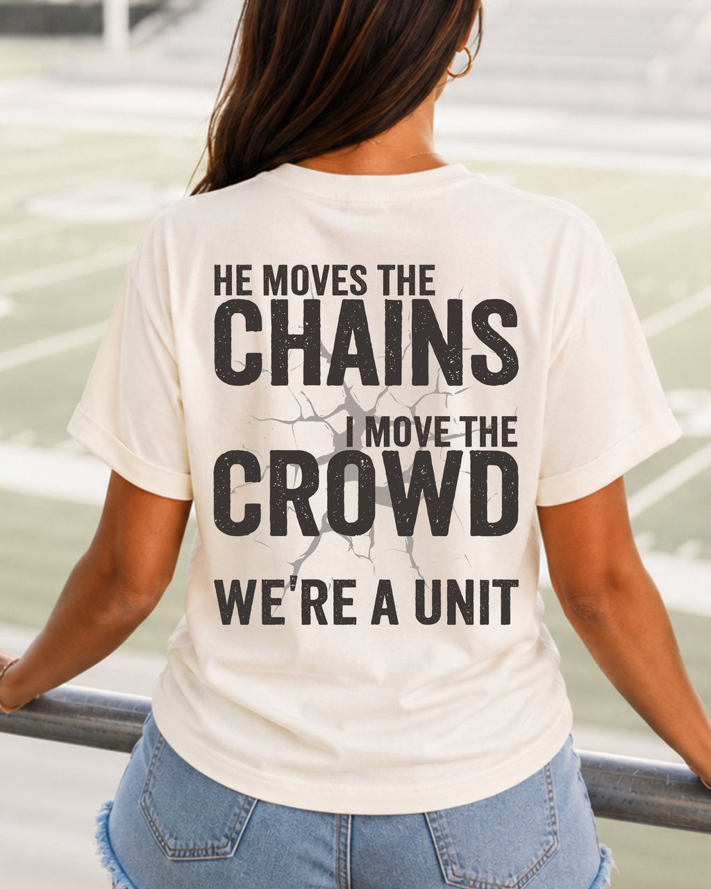 Back view of woman in stadium wearing ivory Comfort Colors football mom game day shirt featuring “He Moves the Chains I Move the Crowd We’re a Unit” bold graphic design. Running Back Mom Shirt Quarterback Mom Shirt. Wide Receiver Mom Shirt.