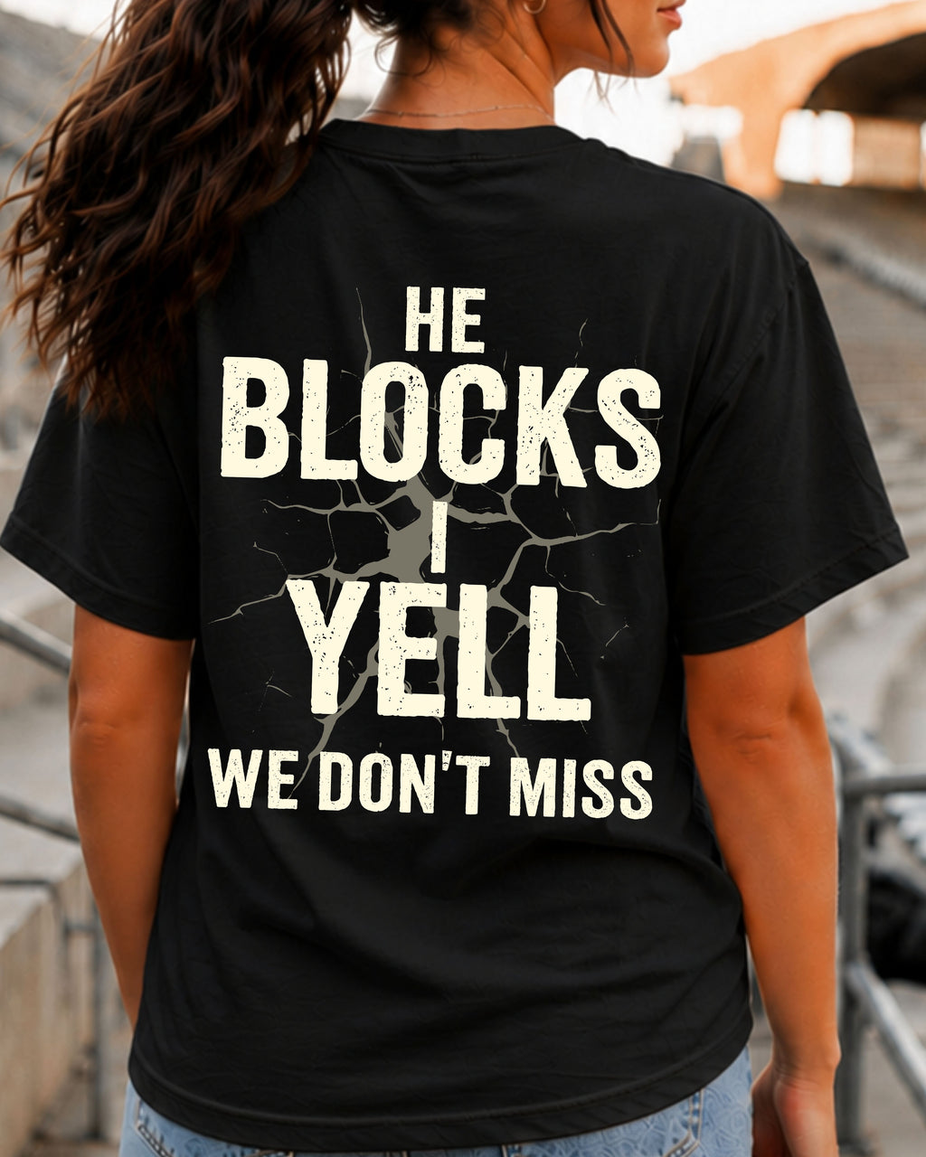 Woman wearing black Comfort Colors football mom game day shirt in stadium with back design that reads “He blocks I yell we don’t miss” in distressed text with cracked graphic