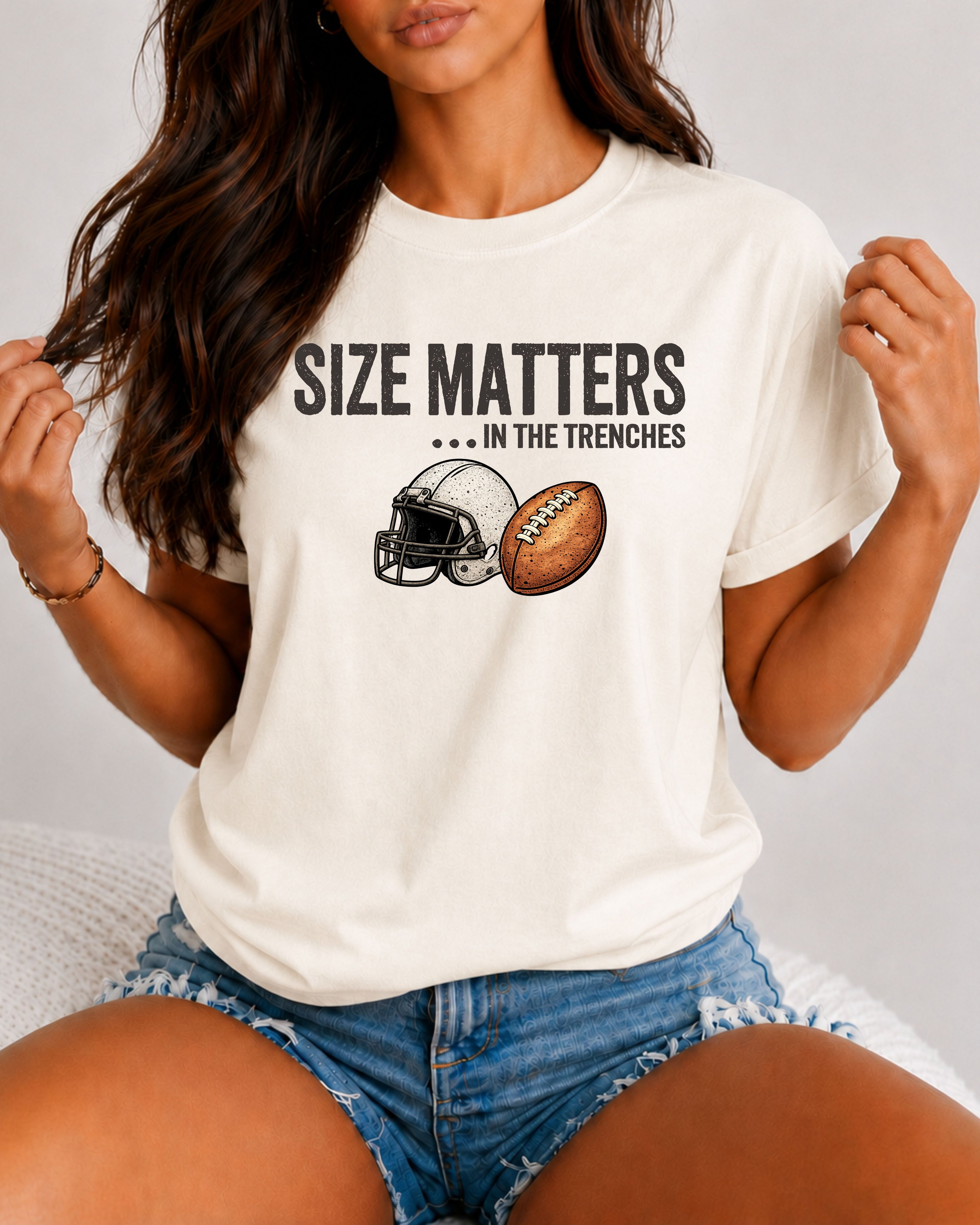 A woman wearing an ivory Comfort Colors t-shirt featuring the funny football saying "Size Matters... In the Trenches" with a vintage football helmet and football graphic. Perfect game day shirt for football fans, offensive linemen, lineman moms, football moms, coaches, players, and anyone who knows games are won in the trenches.