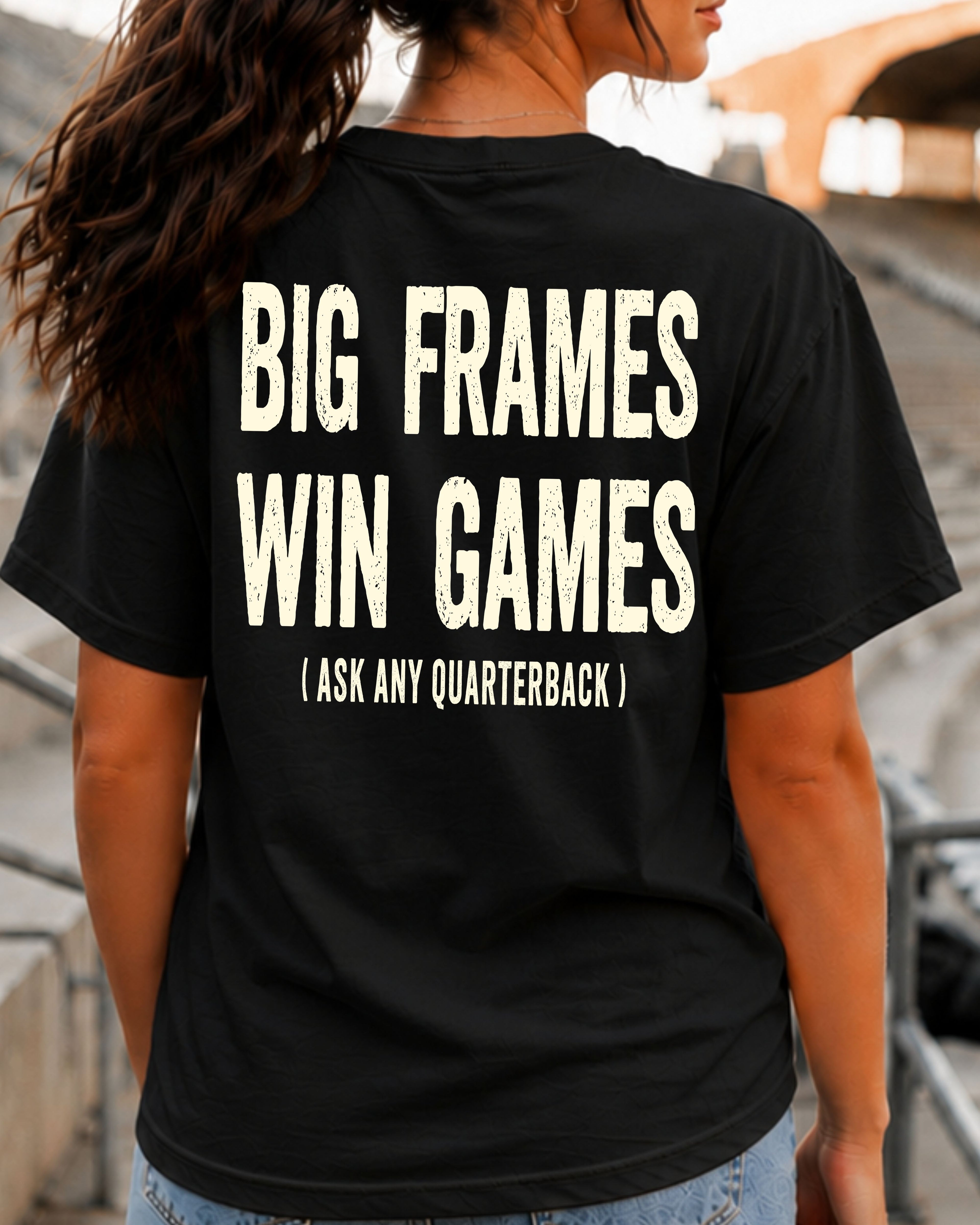 Back view of a woman standing in football stadium bleachers wearing a black Comfort Colors t-shirt with the bold distressed phrase, "BIG FRAMES WIN GAMES (ASK ANY QUARTERBACK)." A relaxed ponytail and blurred stadium background create the perfect game day look for football fans, offensive line parents, and linemen.