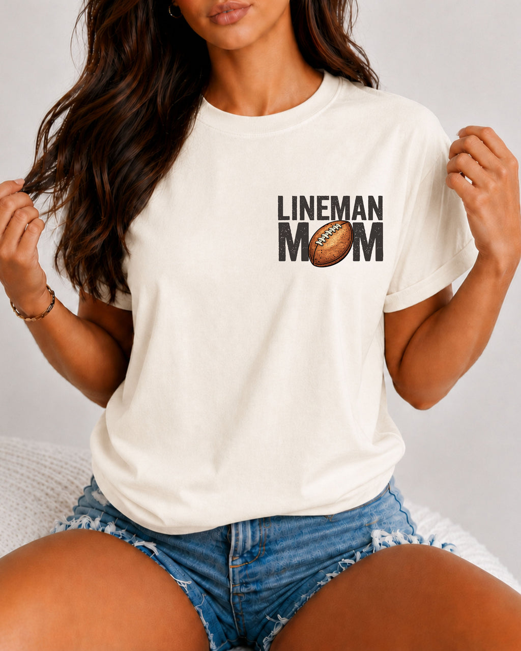 Front view of a woman wearing an ivory Comfort Colors t-shirt with a small left chest "LINEMAN MOM" football graphic. The relaxed-fit shirt is paired with distressed denim shorts, highlighting a casual game day look for proud football moms.