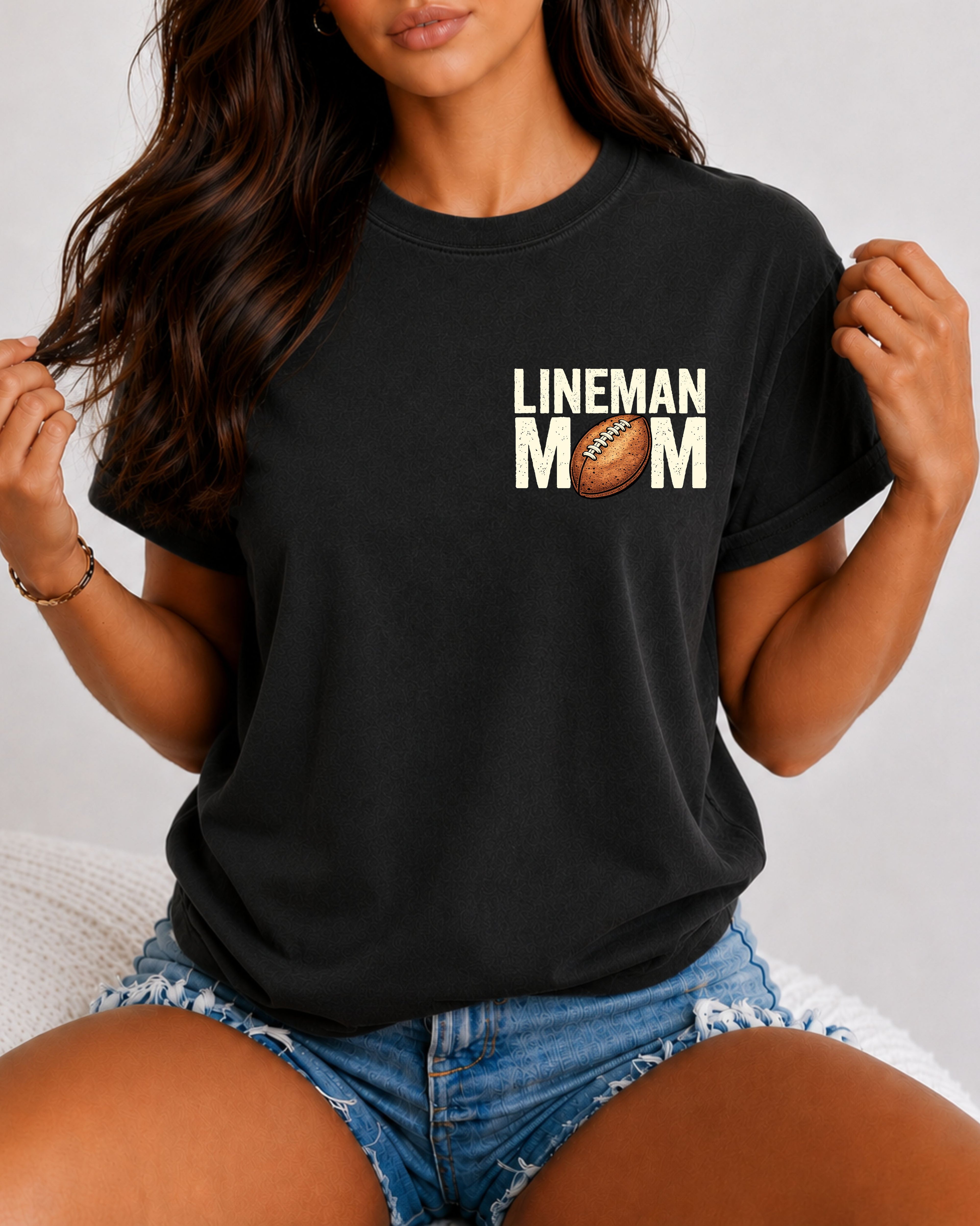 Front view of a woman wearing a black Comfort Colors t-shirt with a small left chest "LINEMAN MOM" football graphic. The relaxed-fit shirt is paired with distressed denim shorts, highlighting a casual game day look for proud football moms.