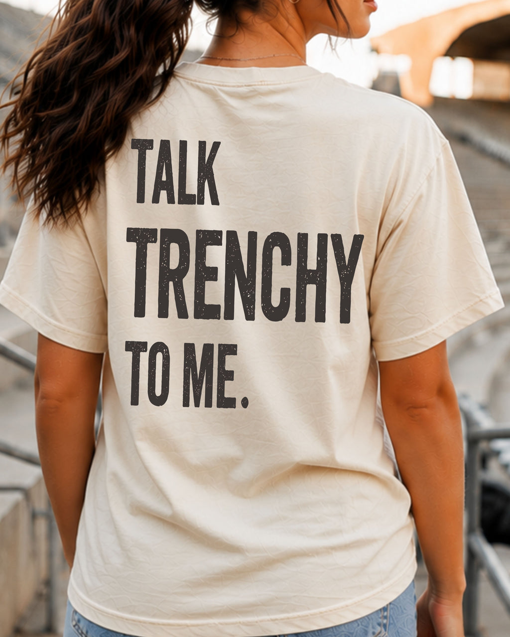 Woman wearing an ivory Comfort Colors football fan T-shirt with the funny back graphic "Talk Trenchy to Me" while standing in stadium bleachers on game day. The oversized graphic is designed for offensive line fans, football moms, dads, and anyone who loves trench football humor.
