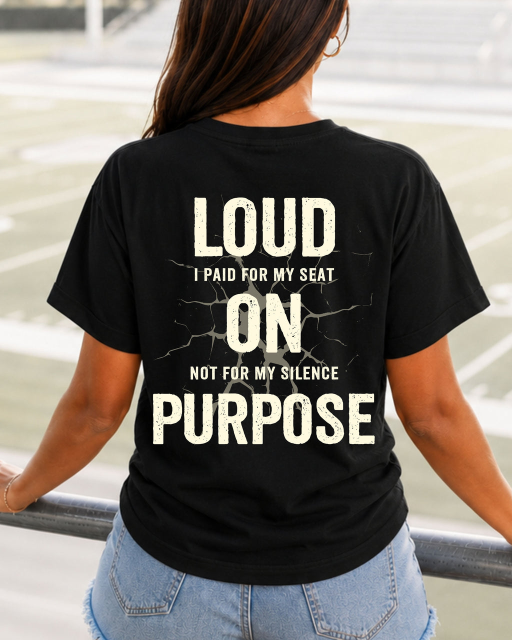 Woman standing in  stadium wearing a black t-shirt with a large distressed cream back graphic that reads “LOUD ON PURPOSE I Paid For My Seat Not For My Silence” with a cracked texture effect. Football Mom Shirt. Lineman Mom Shirt.