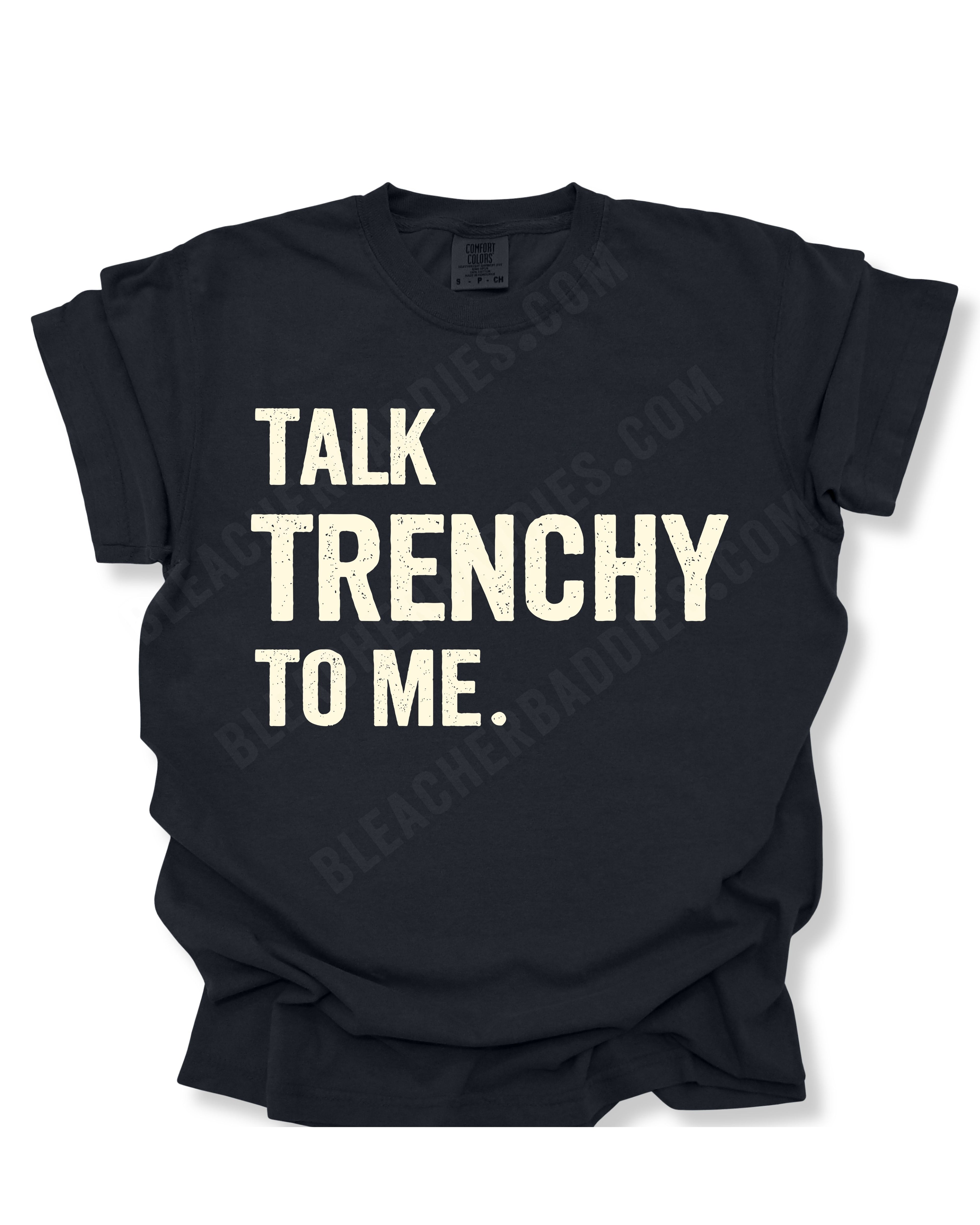 Flat lay of a black Comfort Colors t-shirt featuring the distressed ivory text "TALK TRENCHY TO ME." in large block letters. The shirt is displayed on a white background with rolled sleeves, showcasing a bold football offensive line-inspired graphic perfect for linemen, football players, coaches, and trench football fans.