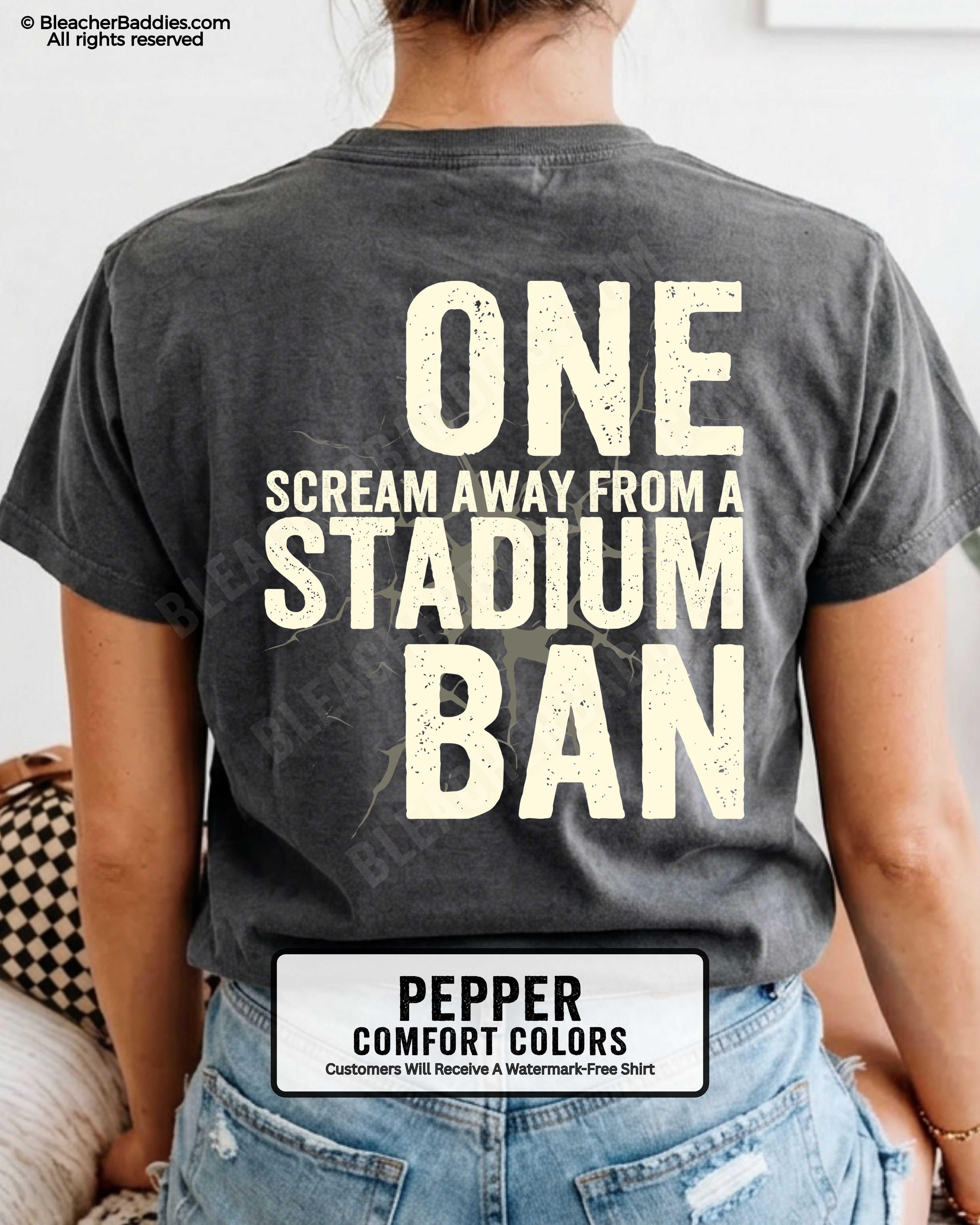 Woman wearing a pepper dark gray  t-shirt with a large distressed cream back graphic that reads “One Scream Away From a Stadium Ban” with a cracked texture effect across the design.