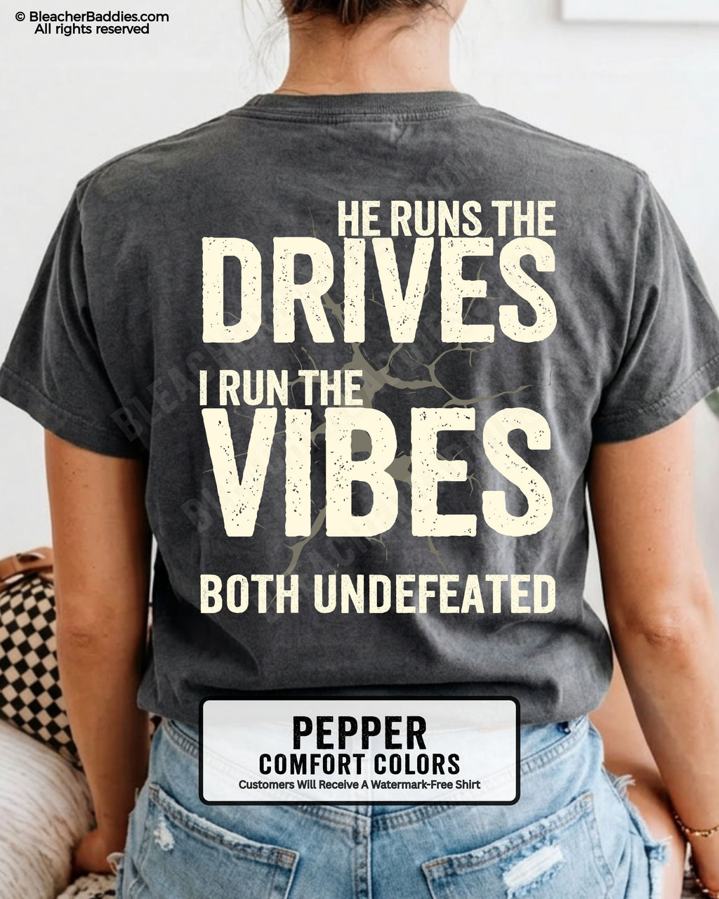 Woman wearing a pepper Comfort Colors football mom shirt from the back featuring the phrase "He Runs the Drives I Run the Vibes Both Undefeated" in distressed Typography with a cracked background graphic.
