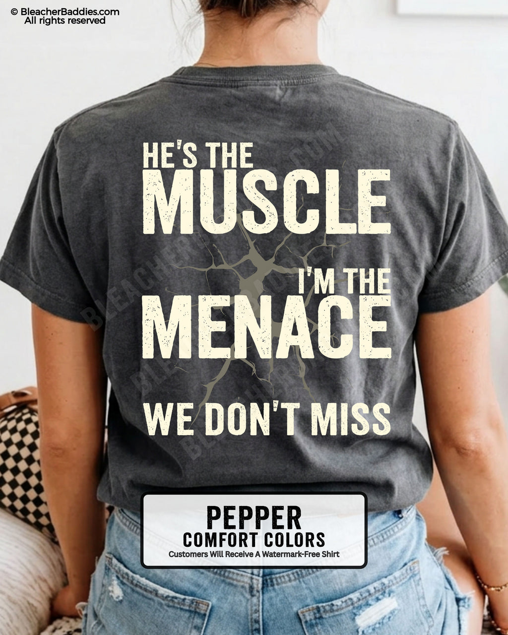 Woman wearing PEPPER Comfort Colors football mom game day shirt with back design that reads “He’s the muscle I’m the menace we don’t miss” in distressed CREAM text with cracked graphic and chambray color label shown