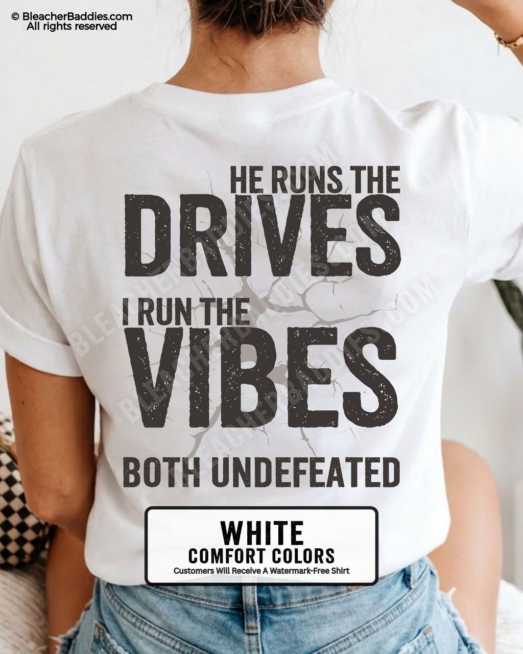 Woman wearing a white Comfort Colors football mom shirt from the back featuring the phrase "He Runs the Drives I Run the Vibes Both Undefeated" in distressed Typography with a cracked background graphic.