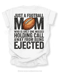 Back of white Comfort Colors football mom shirt with bold graphic text that reads “Just a football mom who is only one missed holding call away from being ejected,” featuring a distressed football design centered on the shirt.