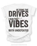 White Comfort Colors football mom shirt featuring the phrase "He Runs the Drives I Run the Vibes Both Undefeated" in distressed black typography with a cracked background graphic.