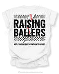 White Comfort Colors Raising Ballers shirt with oversized distressed baseball graphic, black checkerboard stripes, red baseball stitching, and “Not Chasing Participation Trophies” slogan on the back.
