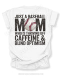 White baseball mom graphic tee featuring distressed black text reading “Just a baseball mom who is thriving off caffeine and blind optimism” with baseball graphic centered in the word MOM