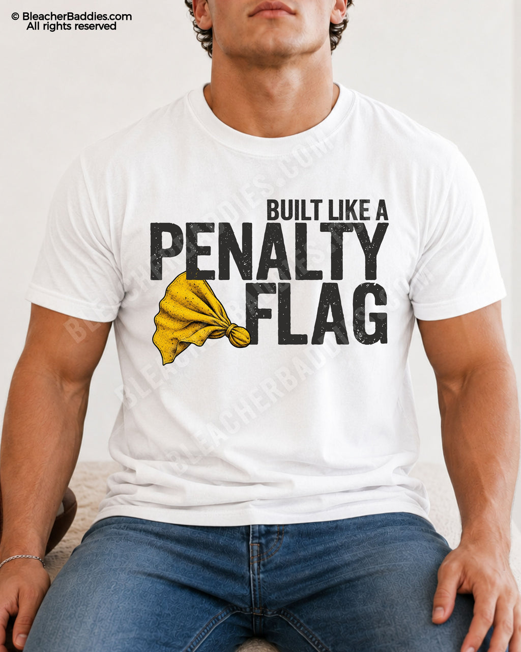 Muscular young male football player model sitting indoors wearing a WHITE Comfort Colors style shirt with distressed “Built Like A Penalty Flag” graphic and yellow penalty flag illustration paired with blue jeans.