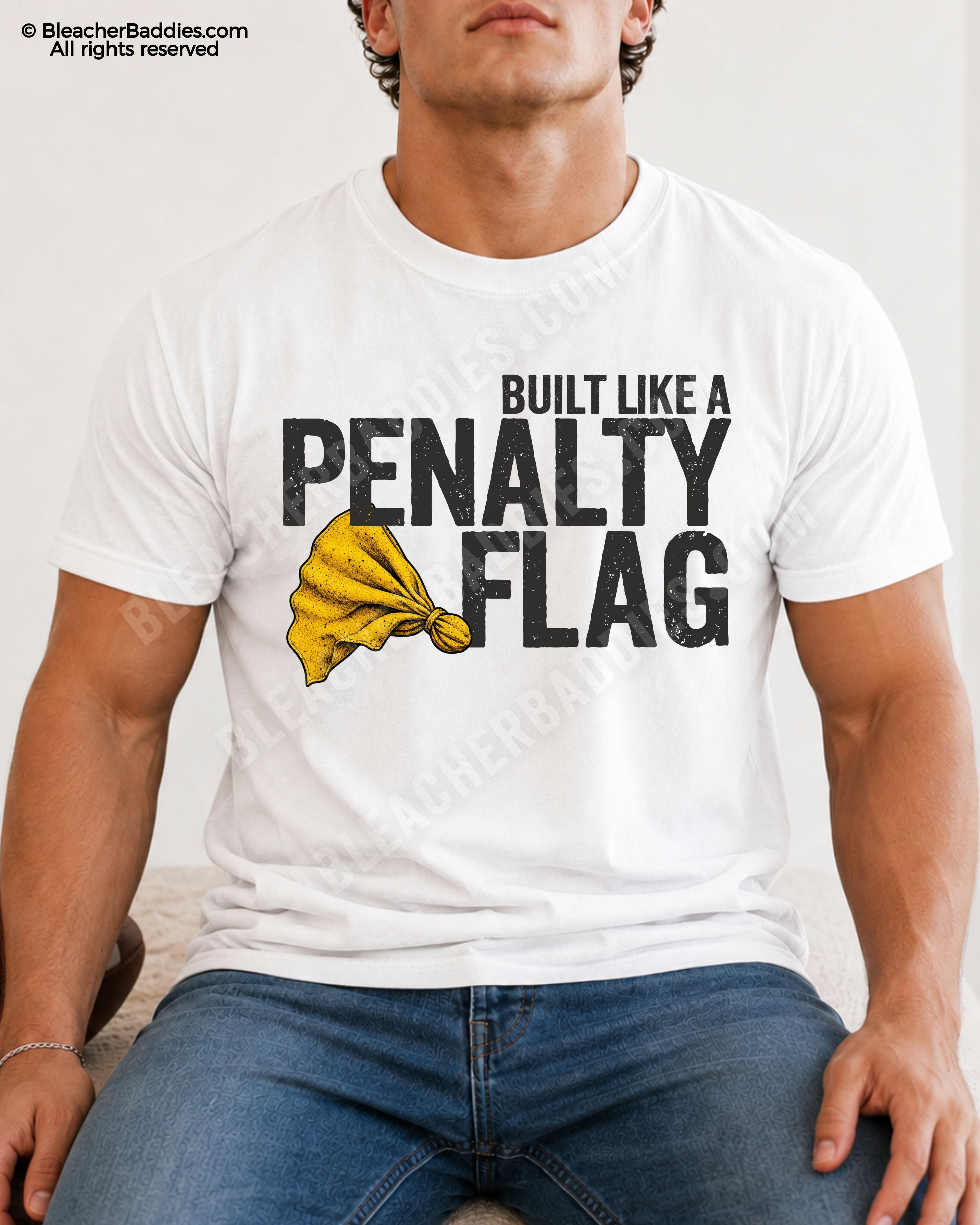 Muscular young male football player model sitting indoors wearing a WHITE Comfort Colors style shirt with distressed “Built Like A Penalty Flag” graphic and yellow penalty flag illustration paired with blue jeans.
