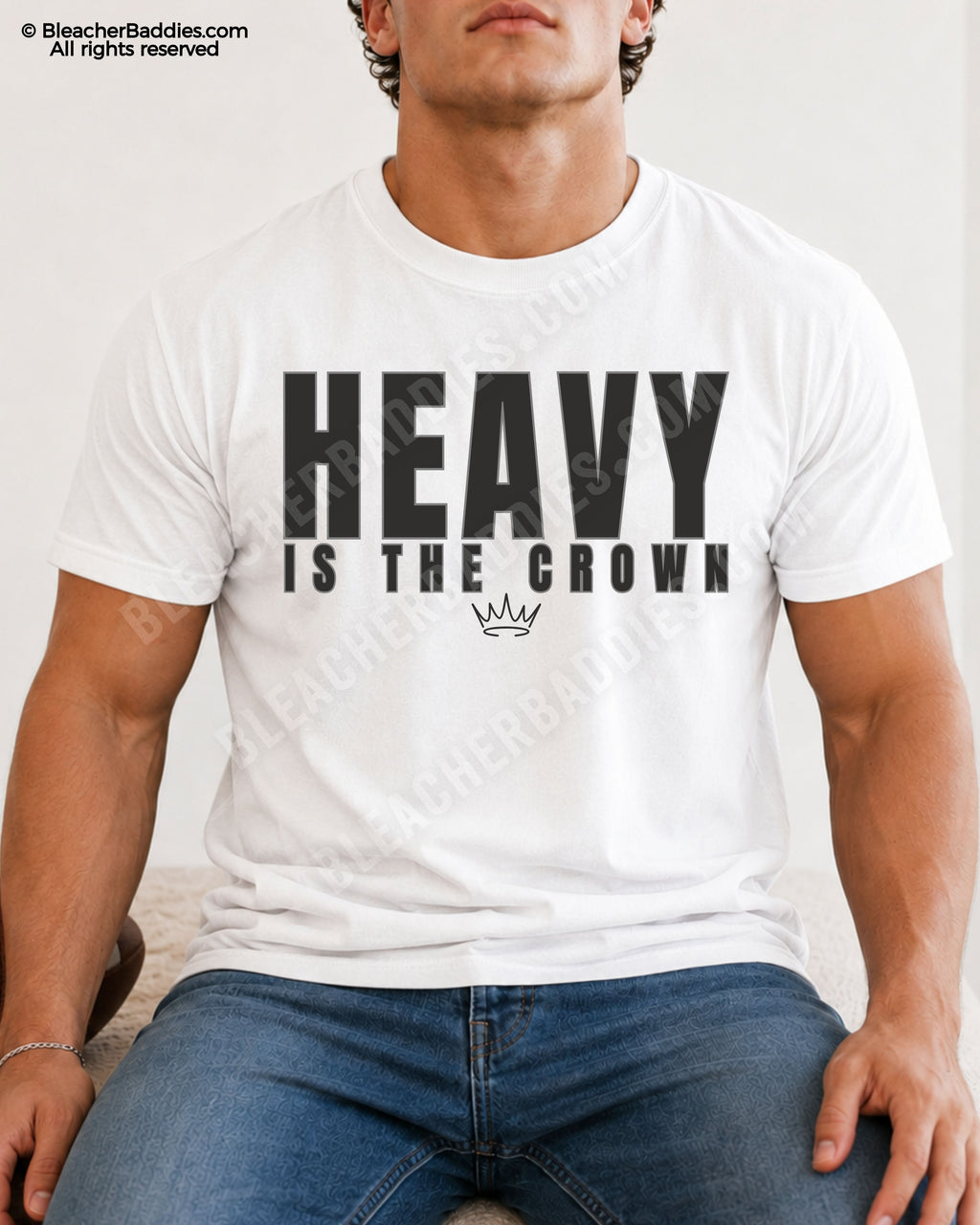 Young male athlete wearing a white “HEAVY IS THE CROWN” motivational football graphic t-shirt while sitting indoors in blue jeans with a relaxed athletic pose.