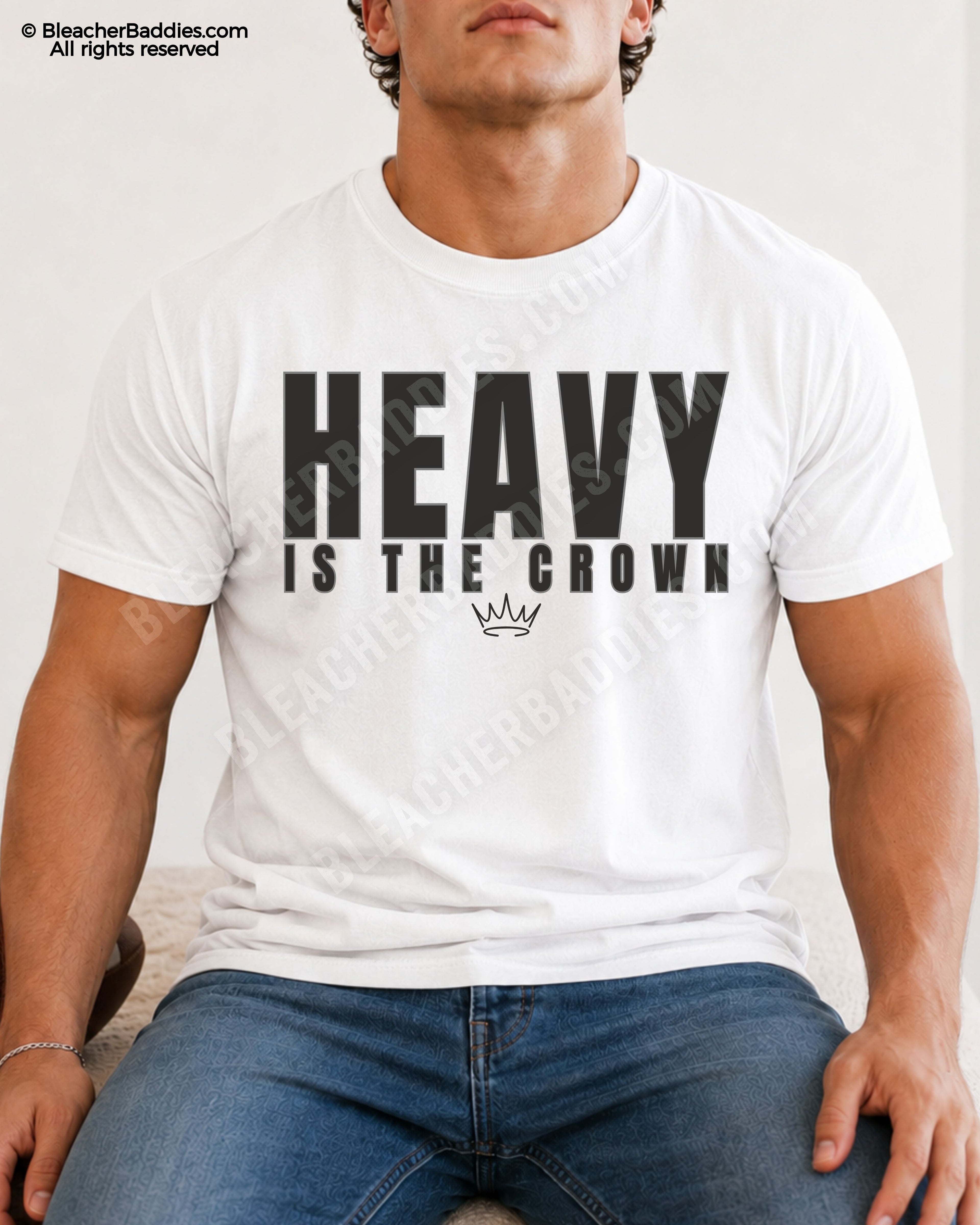 Young male athlete wearing a white “HEAVY IS THE CROWN” motivational football graphic t-shirt while sitting indoors in blue jeans with a relaxed athletic pose.