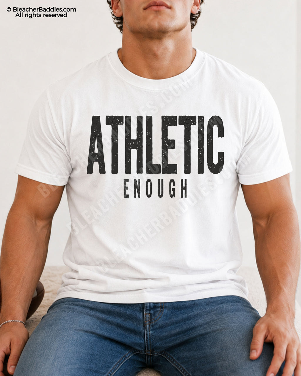 Male football player model wearing a white Comfort Colors “Athletic Enough” football shirt with distressed black typography while seated indoors in jeans.