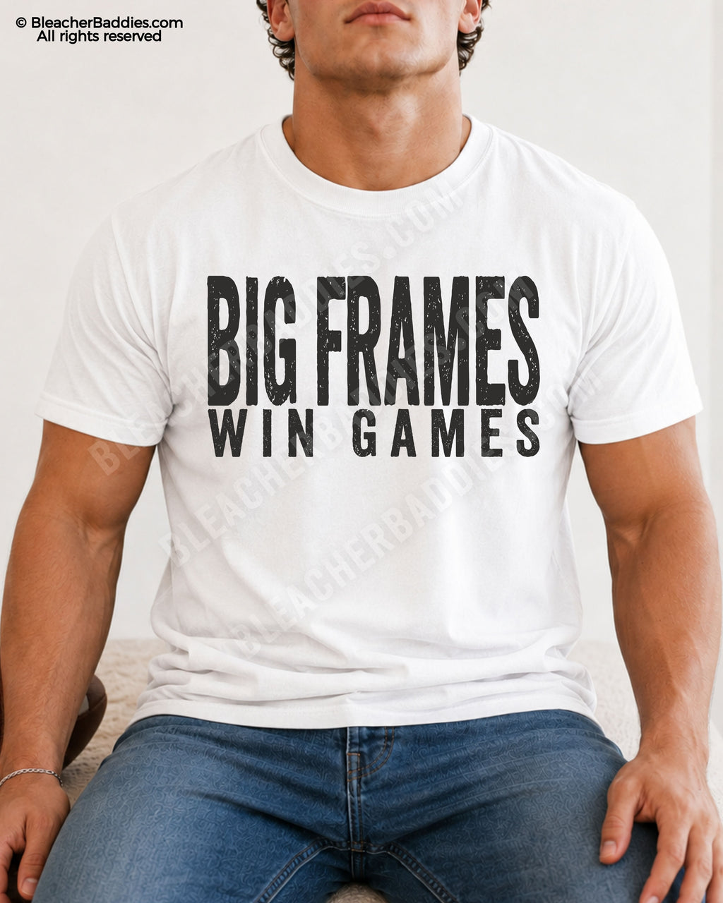 Male football player model wearing a white Comfort Colors “Big Frames Win Games” football shirt with distressed black typography while seated indoors in jeans.