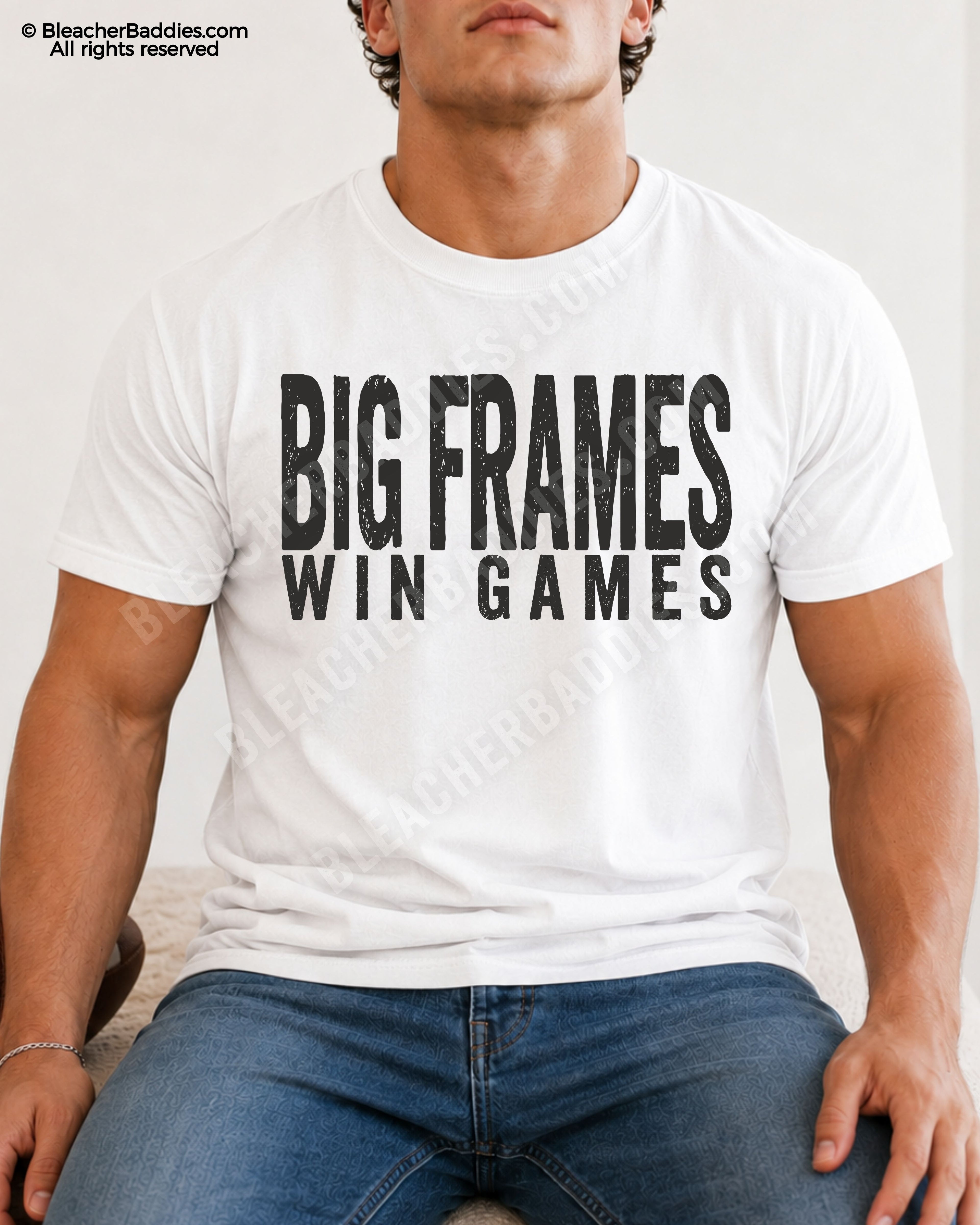 Male football player model wearing a white Comfort Colors “Big Frames Win Games” football shirt with distressed black typography while seated indoors in jeans.