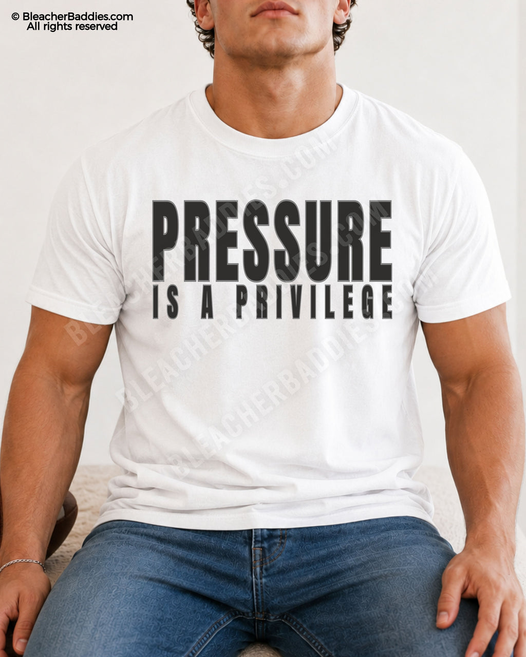 Young male athlete wearing a white motivational football graphic t-shirt with bold black “PRESSURE IS A PRIVILEGE” lettering while sitting indoors in jeans.
