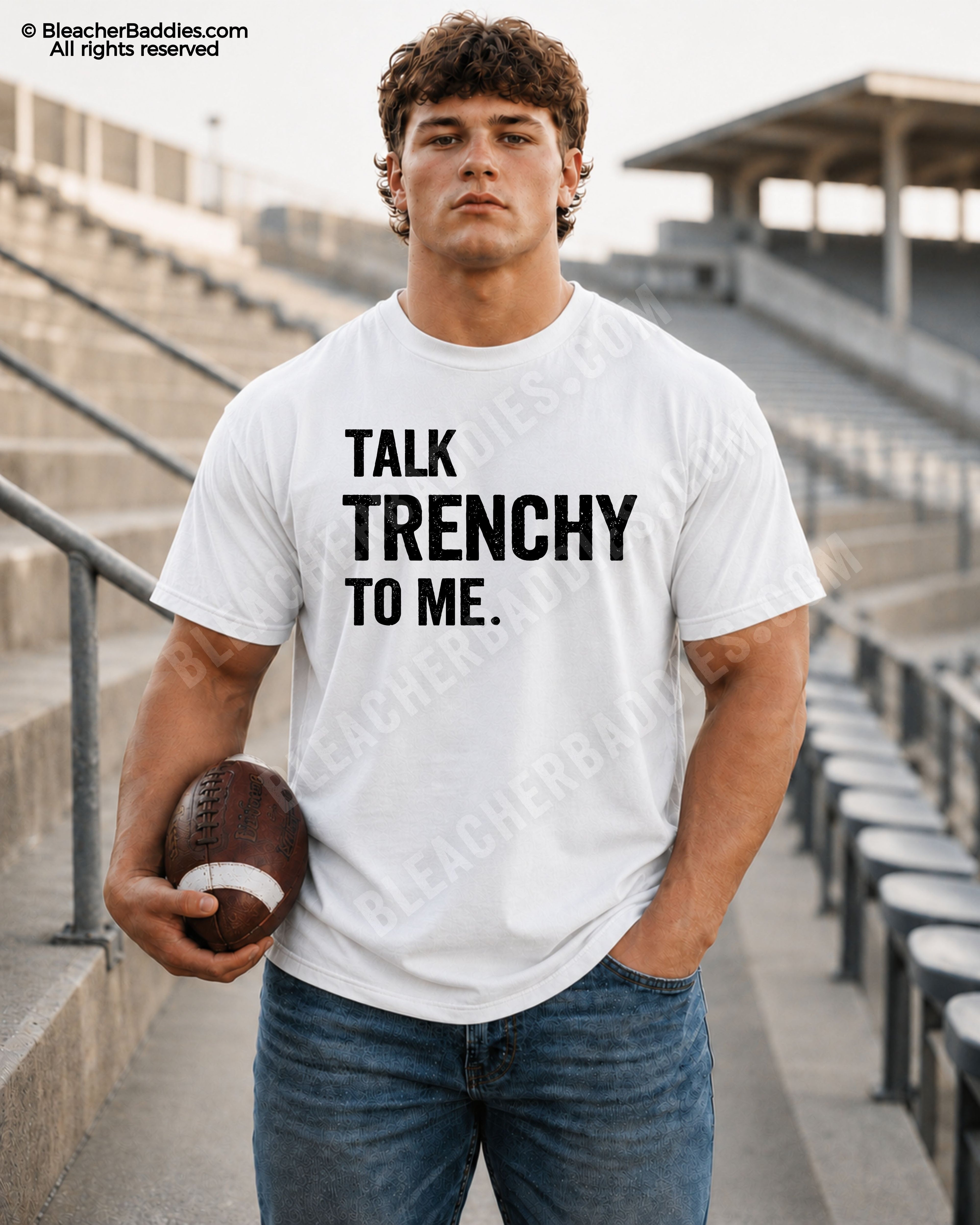 Athletic male model wearing a white Comfort Colors 1717 t-shirt with "Talk Trenchy to Me" graphic, distressed blue jeans, and holding a football in empty stadium bleachers.